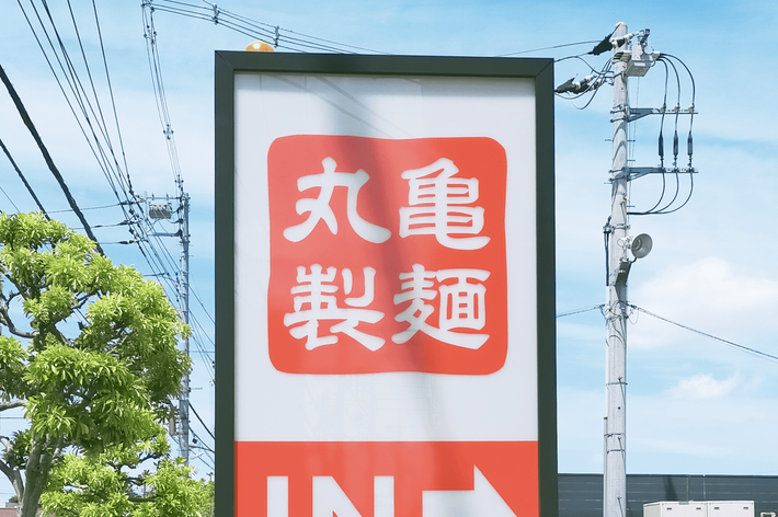 丸亀製麺の看板。赤いロゴと「IN」の矢印が見える。電柱や木が背景にある。