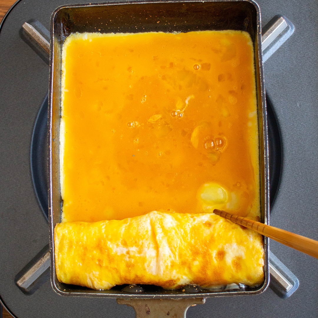 四角いフライパンで作られている卵焼きの工程。巻かれている卵部分に新しい卵液が注がれている。駒倒し調理中。