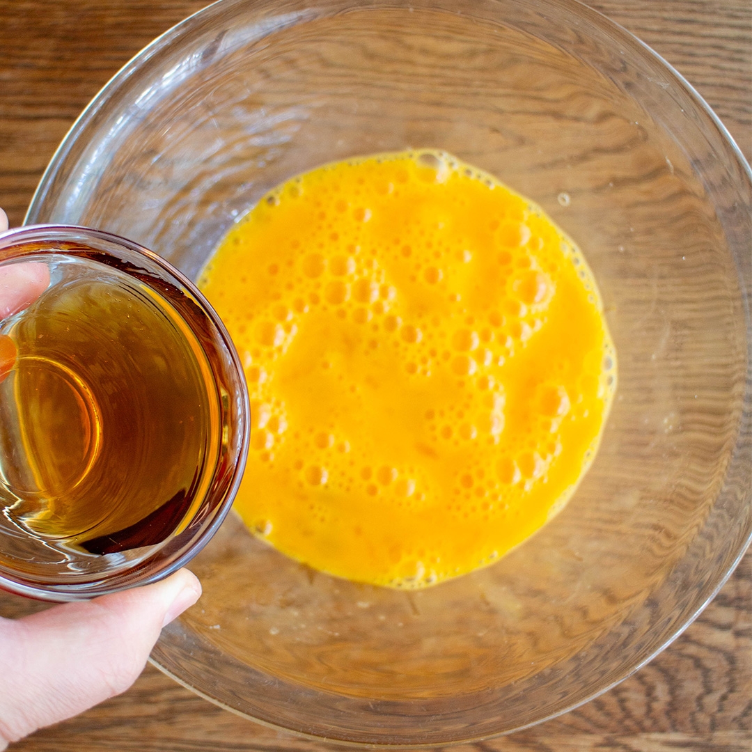 透明なボウルに溶き卵を入れ、別の容器から液体を注ごうとしている手。