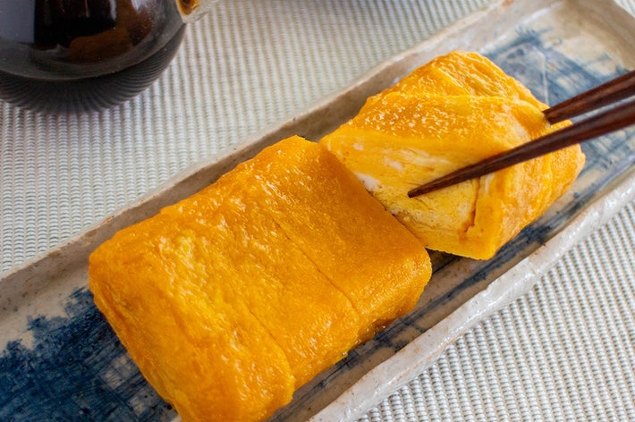 だし巻き卵が陶器の皿に切り分けられ、箸で一切れが持ち上げられている。背景に醤油入れが見える。