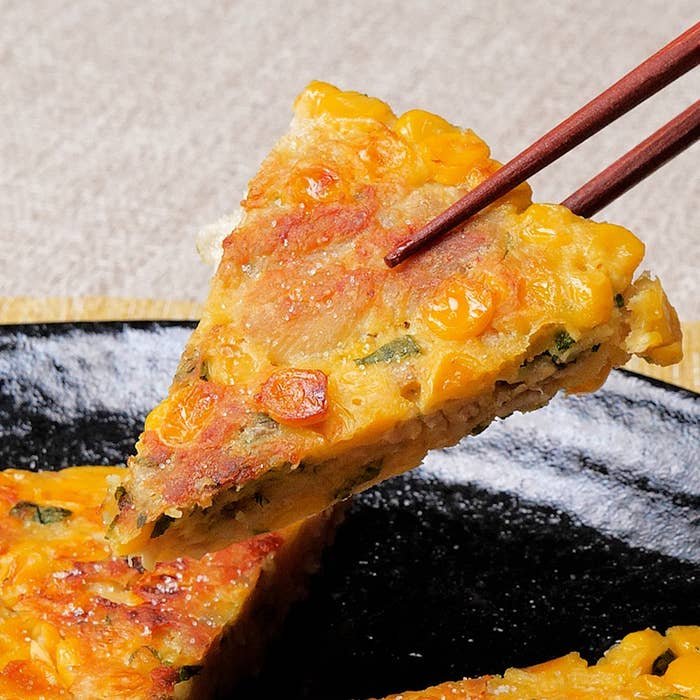 箸で持ち上げられたチヂミの一切れ、トウモロコシやネギが散りばめられた韓国のお好み焼き。