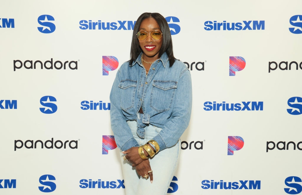 Person in denim shirt and pants stands on SiriusXM and Pandora event backdrop, smiling and wearing large bracelets and glasses