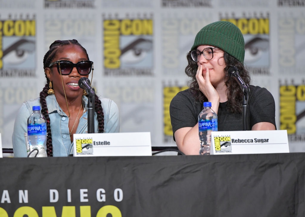 Two people at a Comic-Con panel. One wears sunglasses and braids; the other has long hair and a beanie. They're speaking into microphones