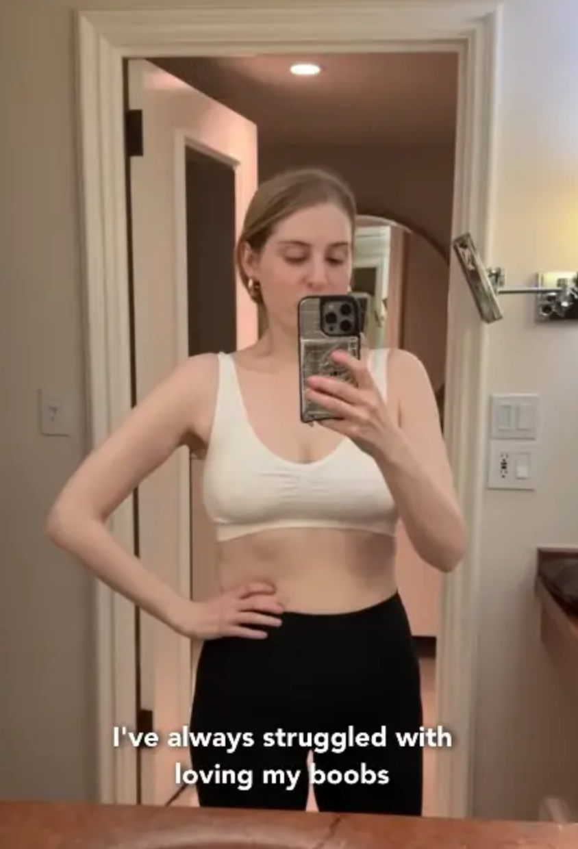 Meghan Trainor in a white sports bra and black pants, taking a mirror selfie in a bathroom. Text below reads: "I've always struggled with loving my boobs."
