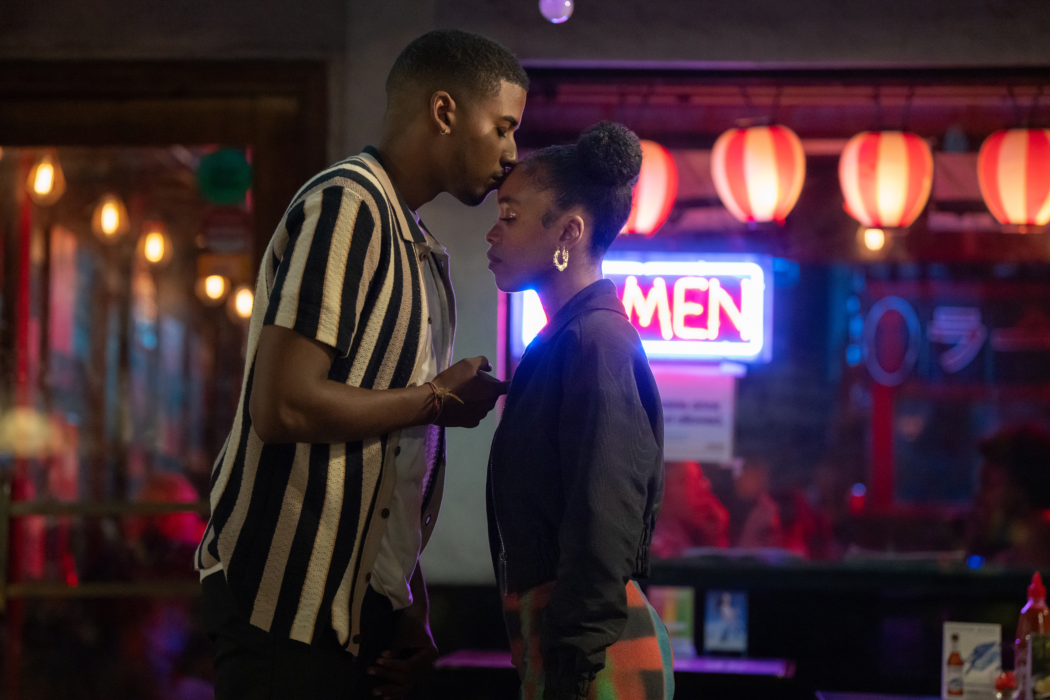 A man kisses a woman's forehead in a warmly lit restaurant decorated with paper lanterns and a vibrant "Ramen" sign