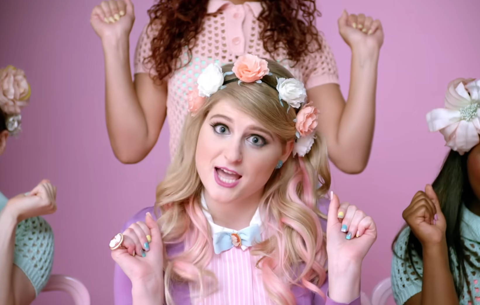 Meghan Trainor in a floral headband and pastel outfit, performing with backup dancers making hand gestures in the "All About That Bass" music video