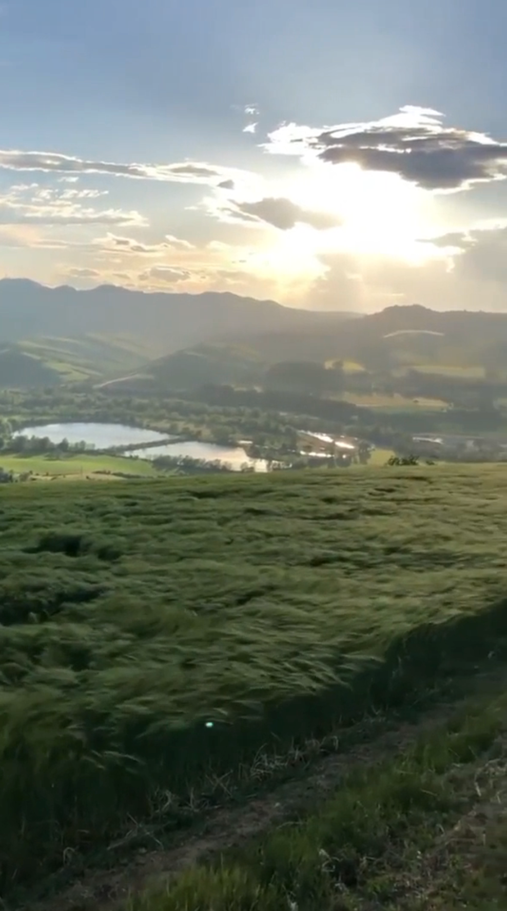 Scenic landscape with rolling hills, a reflective lake, and sun rays breaking through clouds over distant mountains