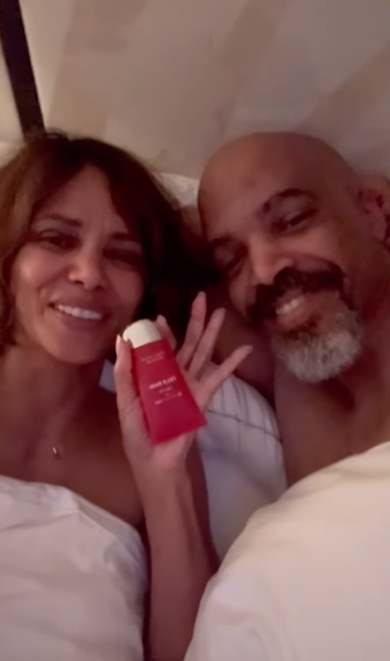 Halle Berry and Van Hunt lying in bed, with Halle holding a bottle of intimacy gel and smiling