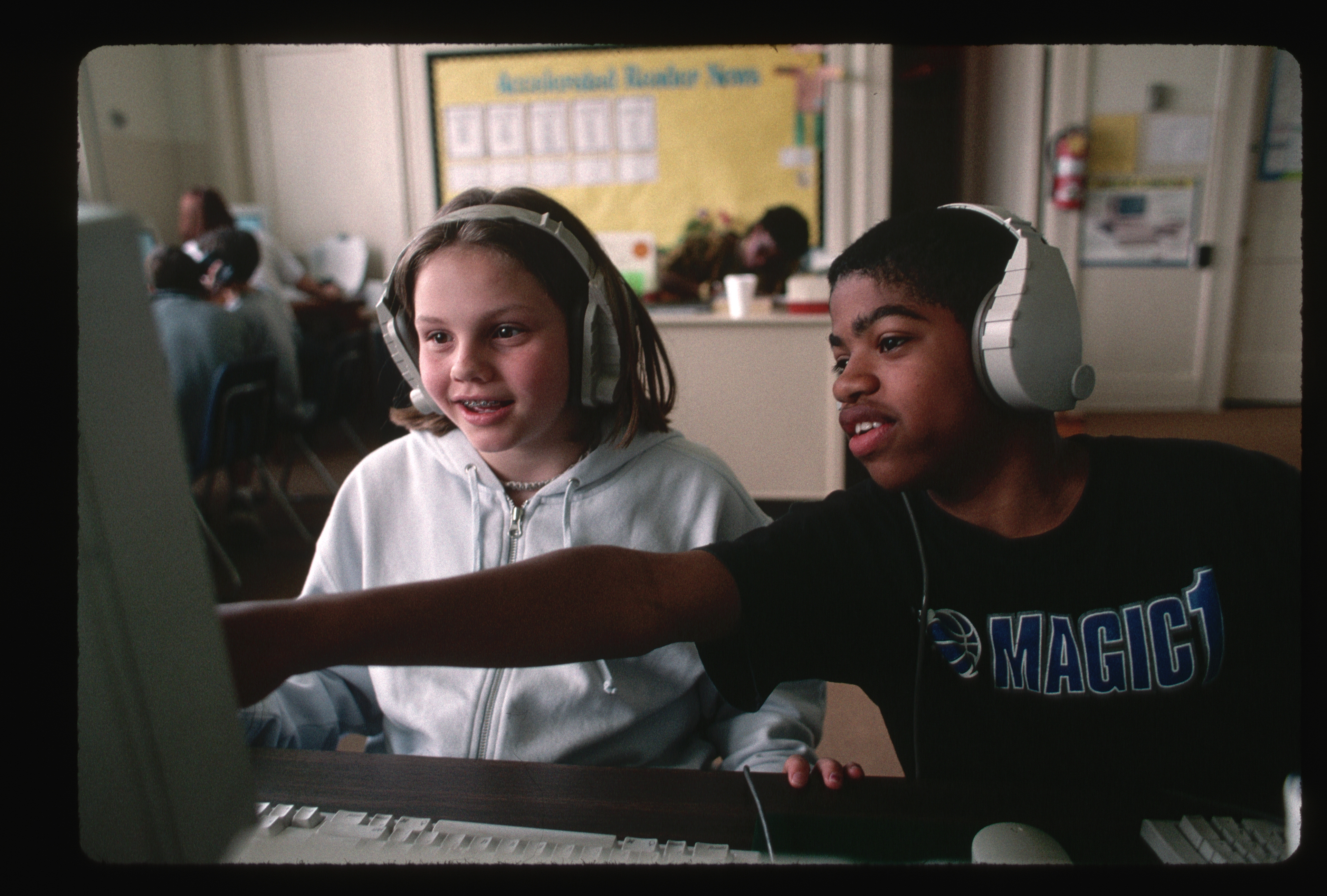 Two students with headphones share a computer, focused and engaged, in a classroom setting