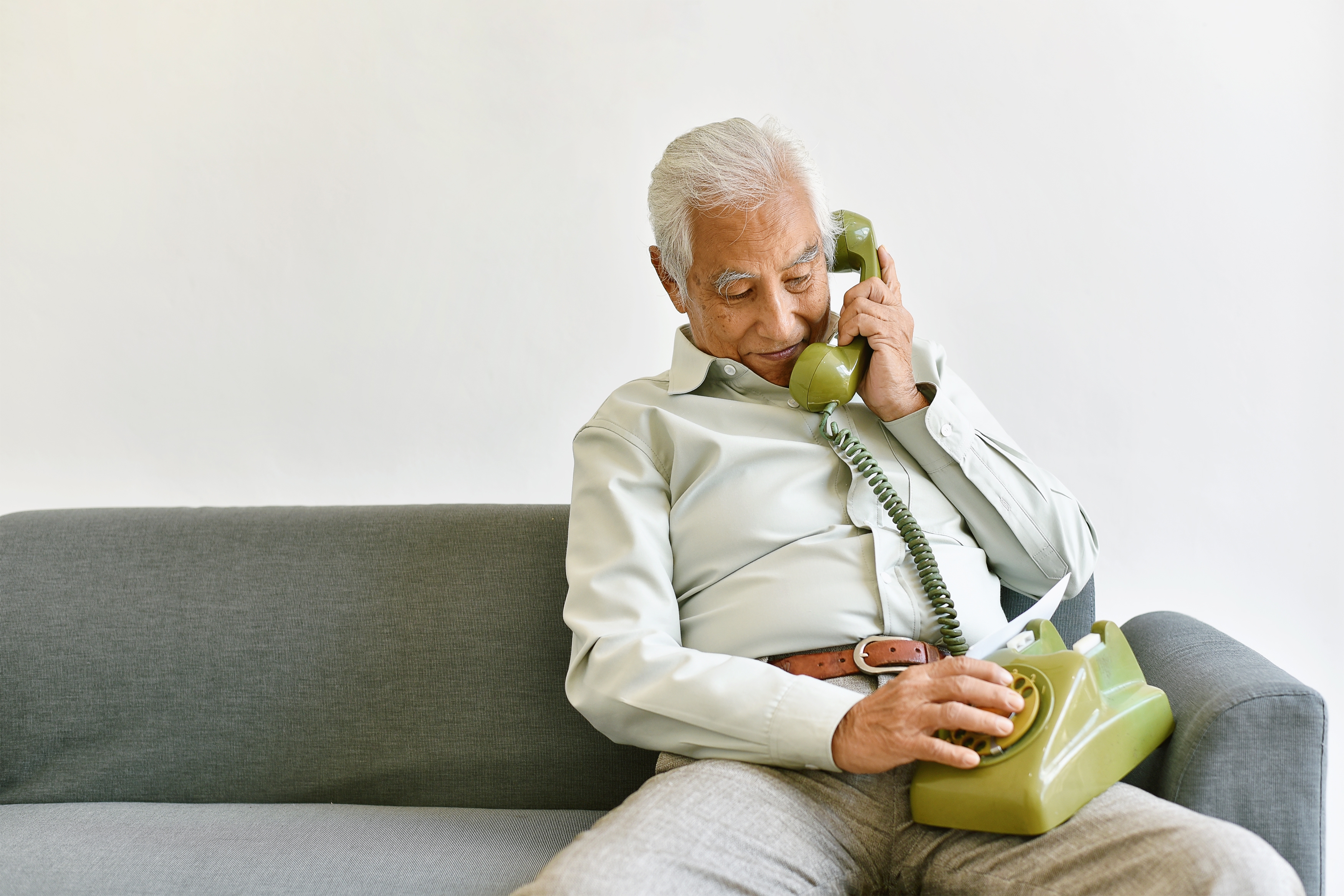 Elderly man sitting on a sofa, cheerfully talking and dialing numbers on a vintage rotary phone