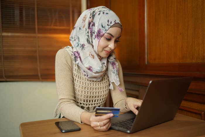 A person in a floral hijab shops online, holding a credit card and using a laptop, with a smartphone nearby on a wooden table