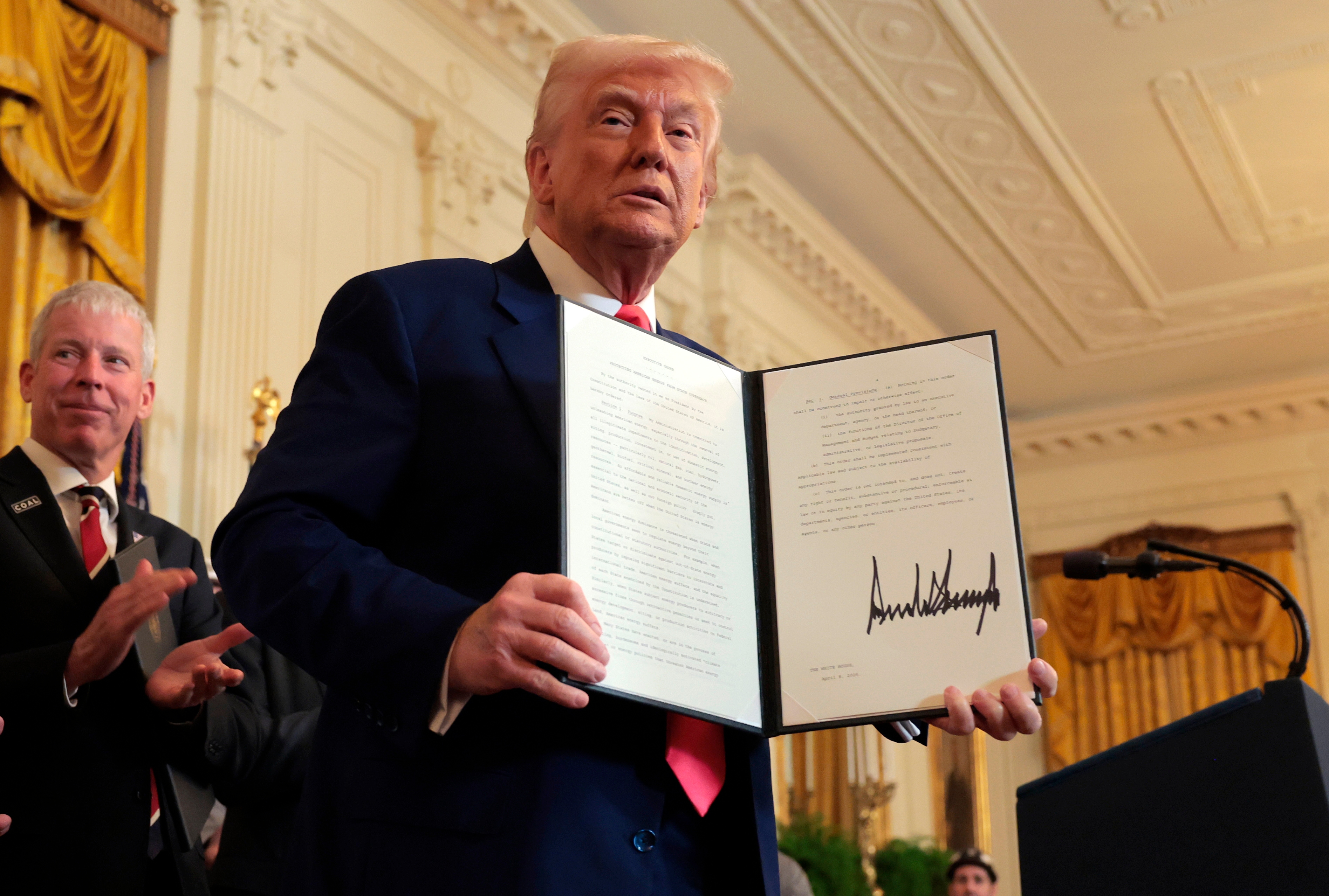 A man in a suit holds up a signed document while standing in front of a crowd in a formal room