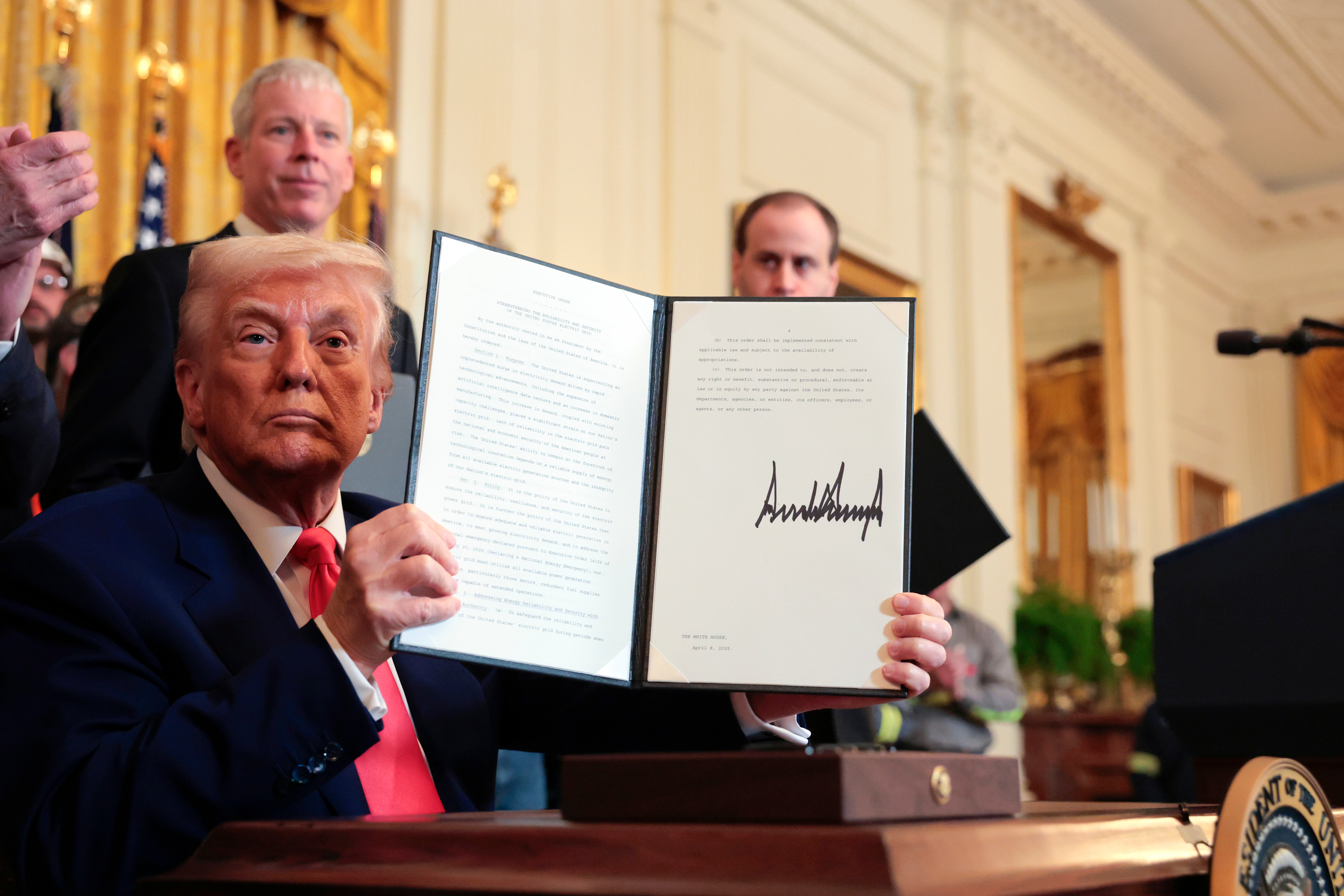 Person seated, holding up a signed document ceremoniously at a formal event, with onlookers in the background