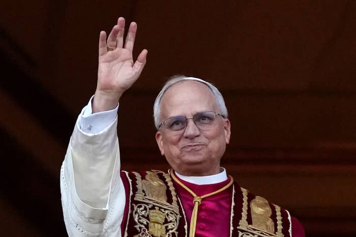 A person in ornate religious attire waves, wearing glasses and a ceremonial robe with intricate patterns