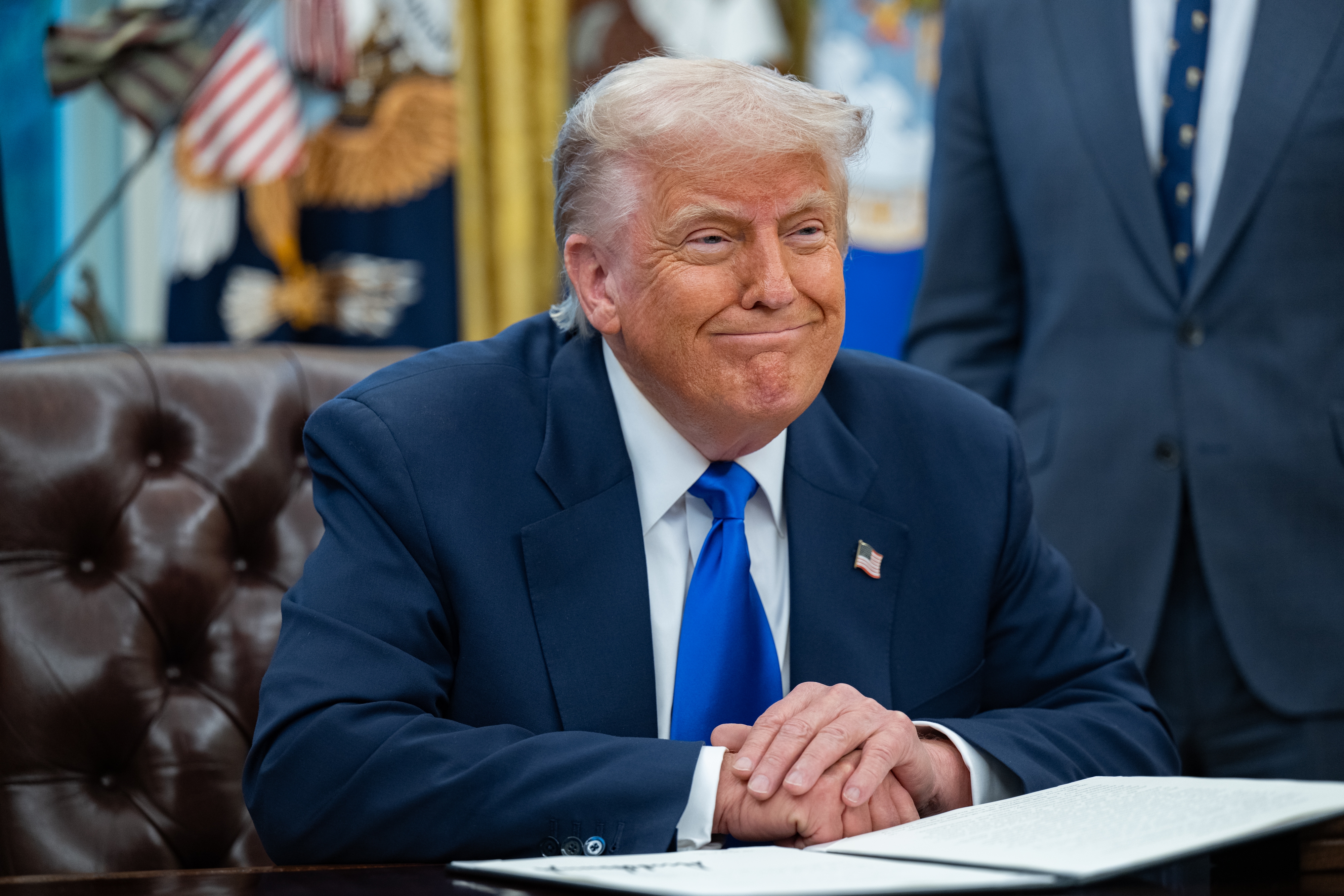 A person sitting at a desk with hands clasped, wearing a suit and blue tie, in an office setting with U.S. flag visible in the background