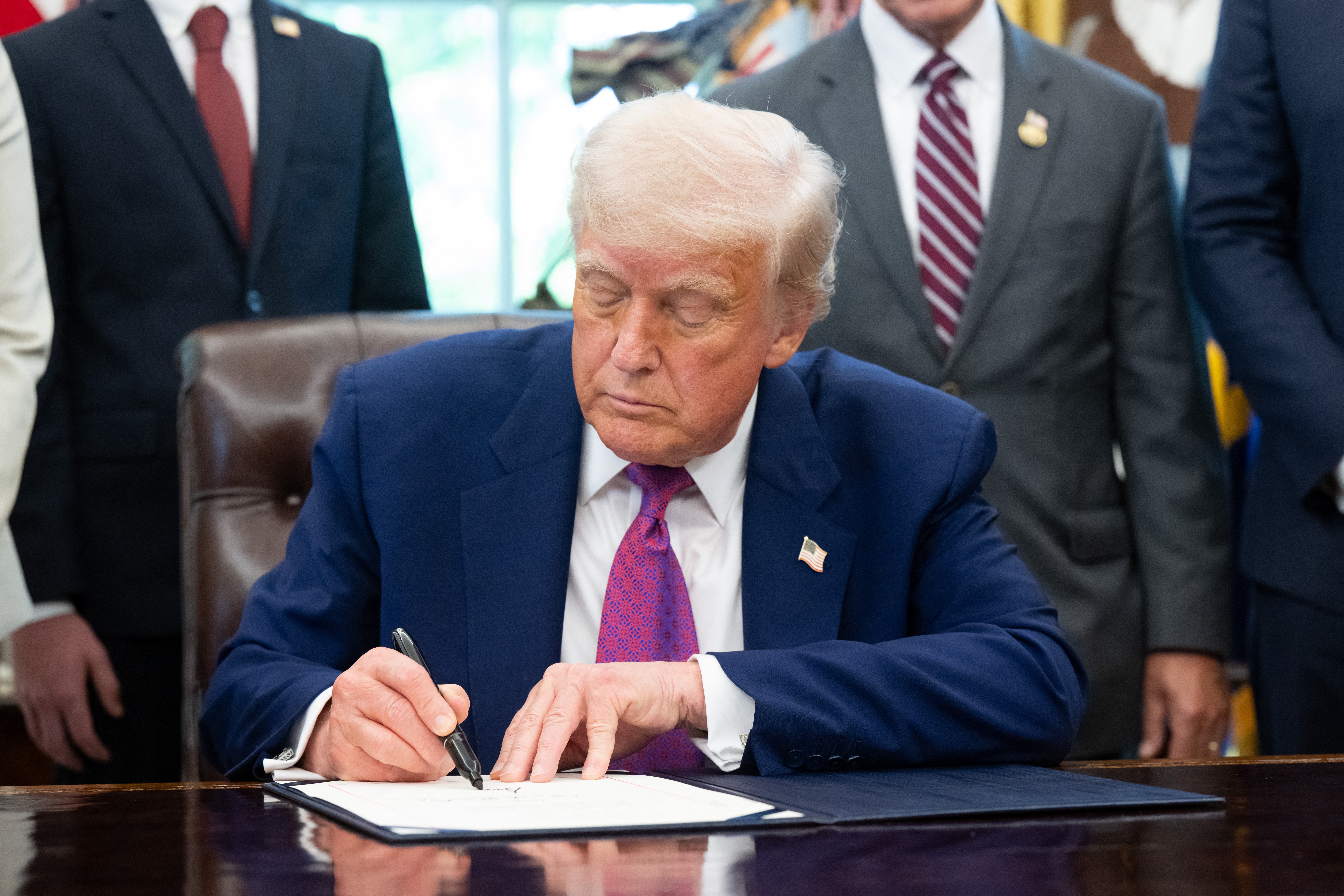 A person signs a document at a desk, surrounded by others in suits, indicating a formal or official setting