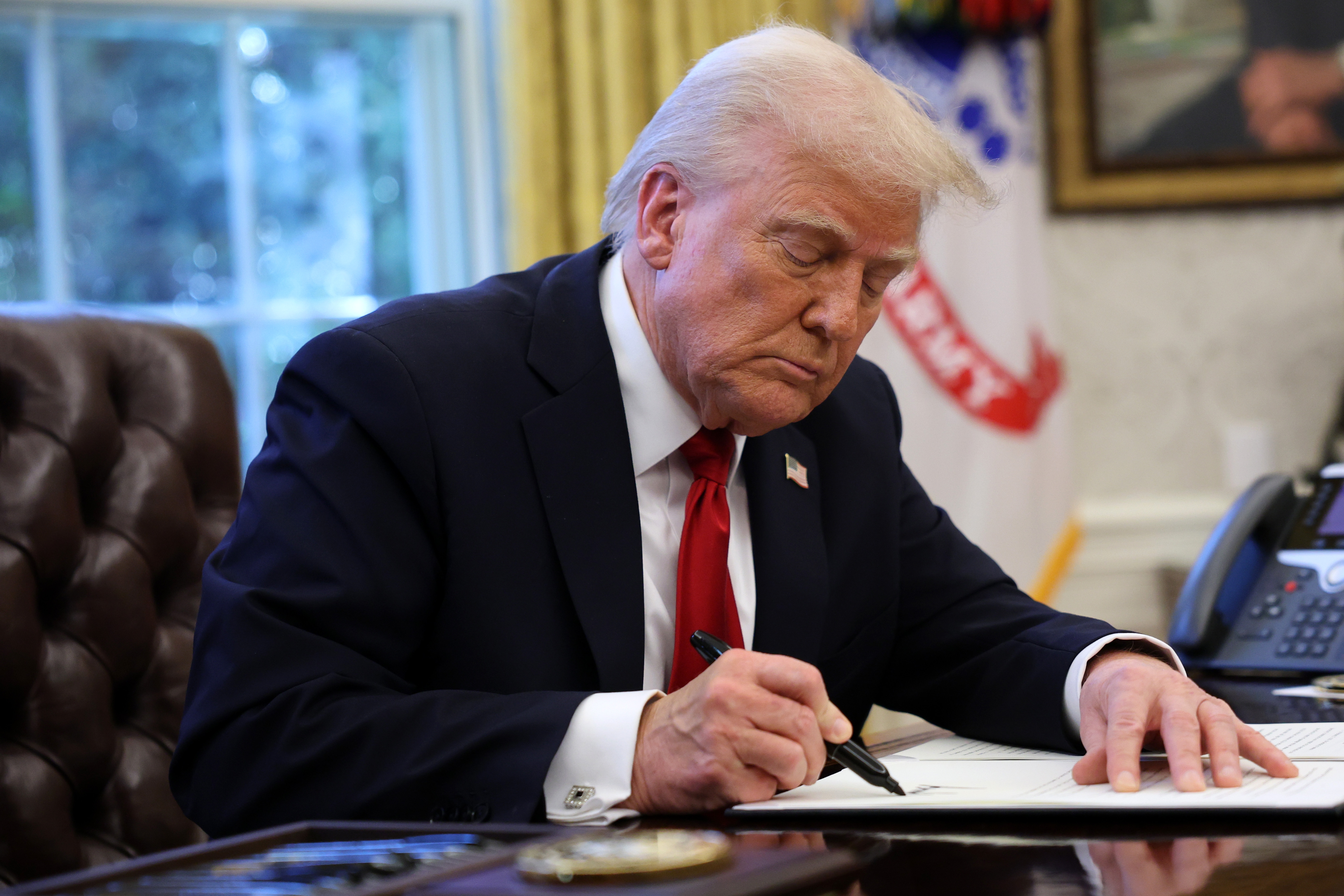 A person in a suit sits at a desk in an office, signing a document with a focused expression