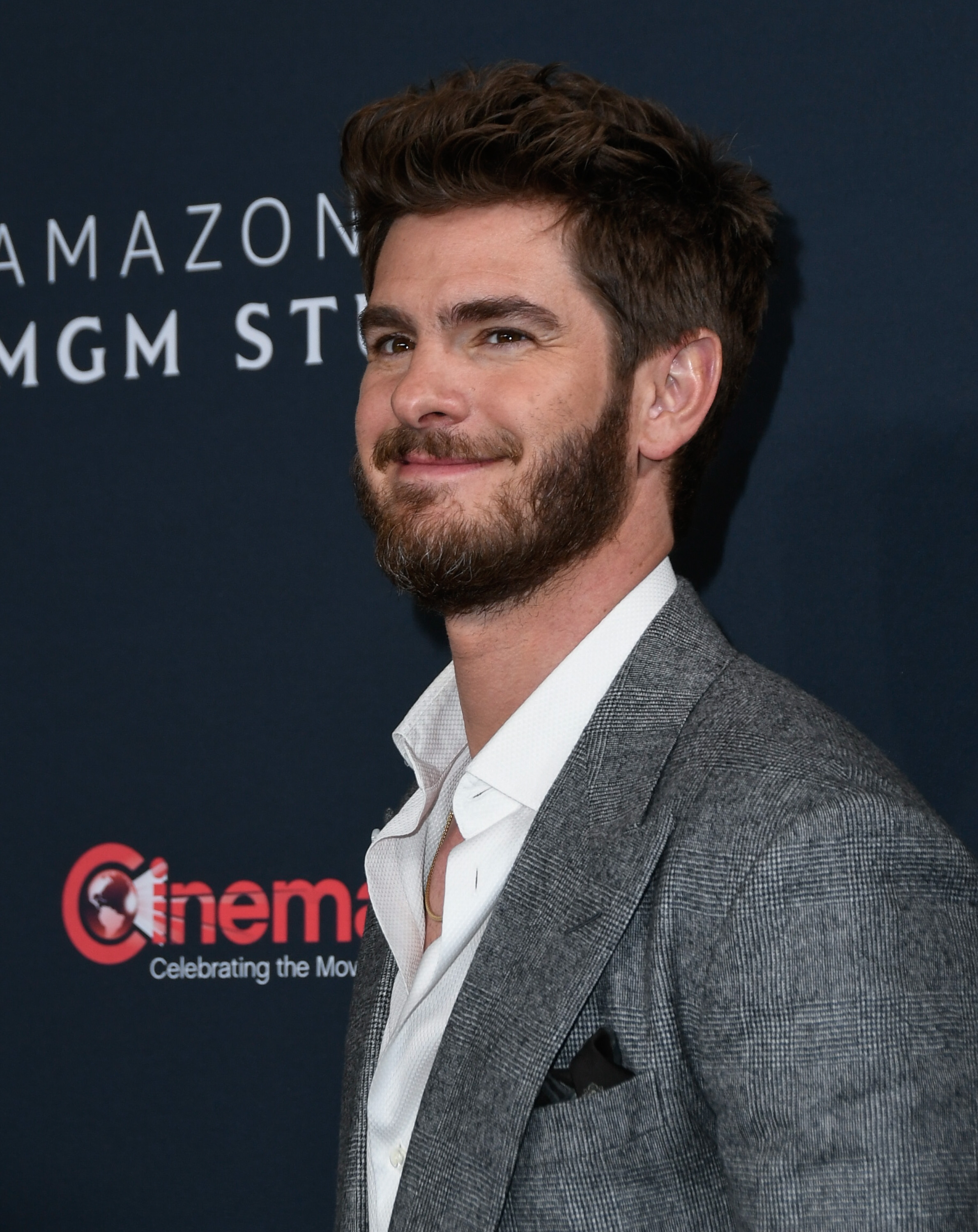 Person smiling at a red carpet event, wearing a gray suit jacket and white shirt