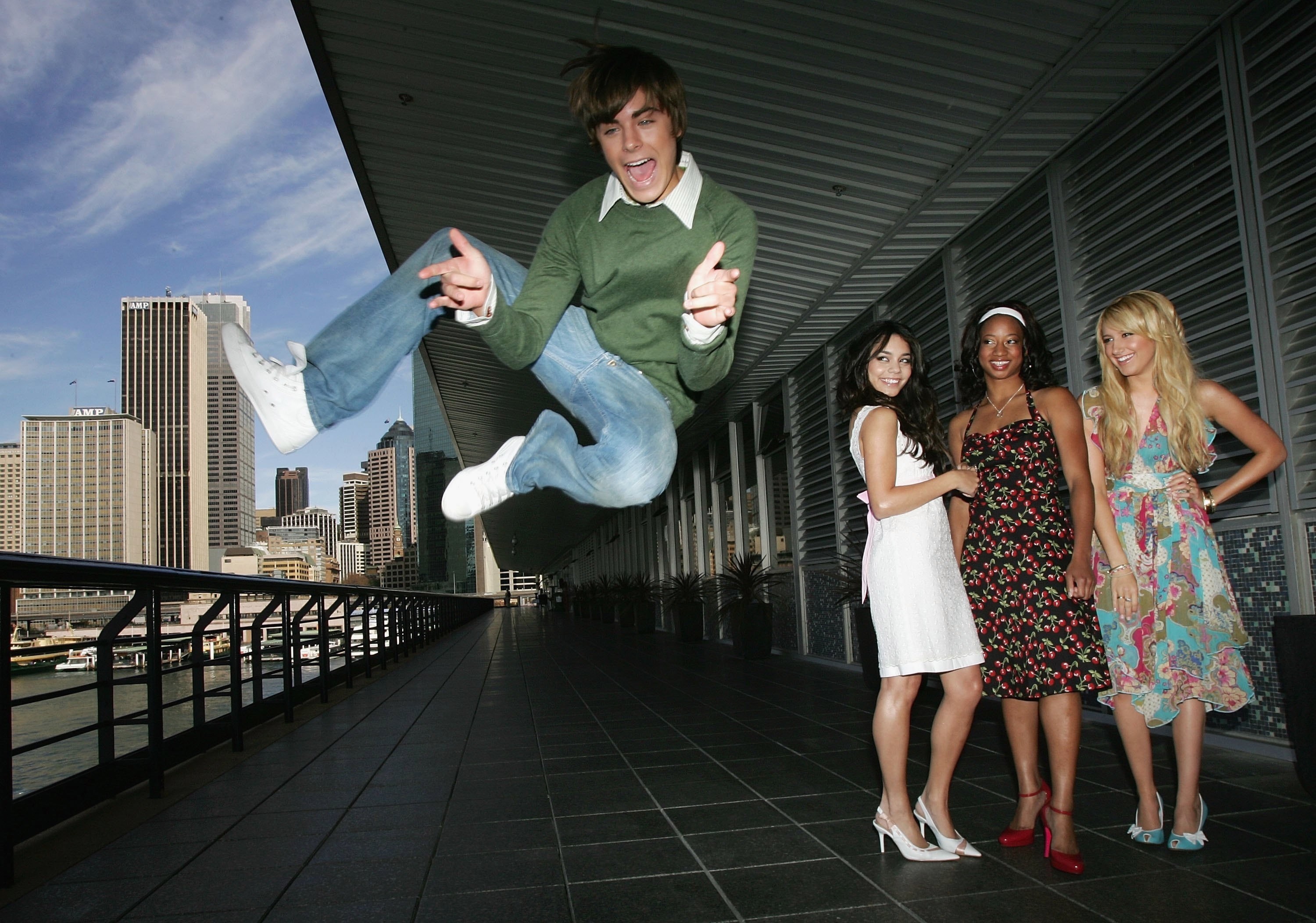 A group of four people posing playfully in an urban setting. One is jumping energetically, others stand smiling in stylish, casual outfits