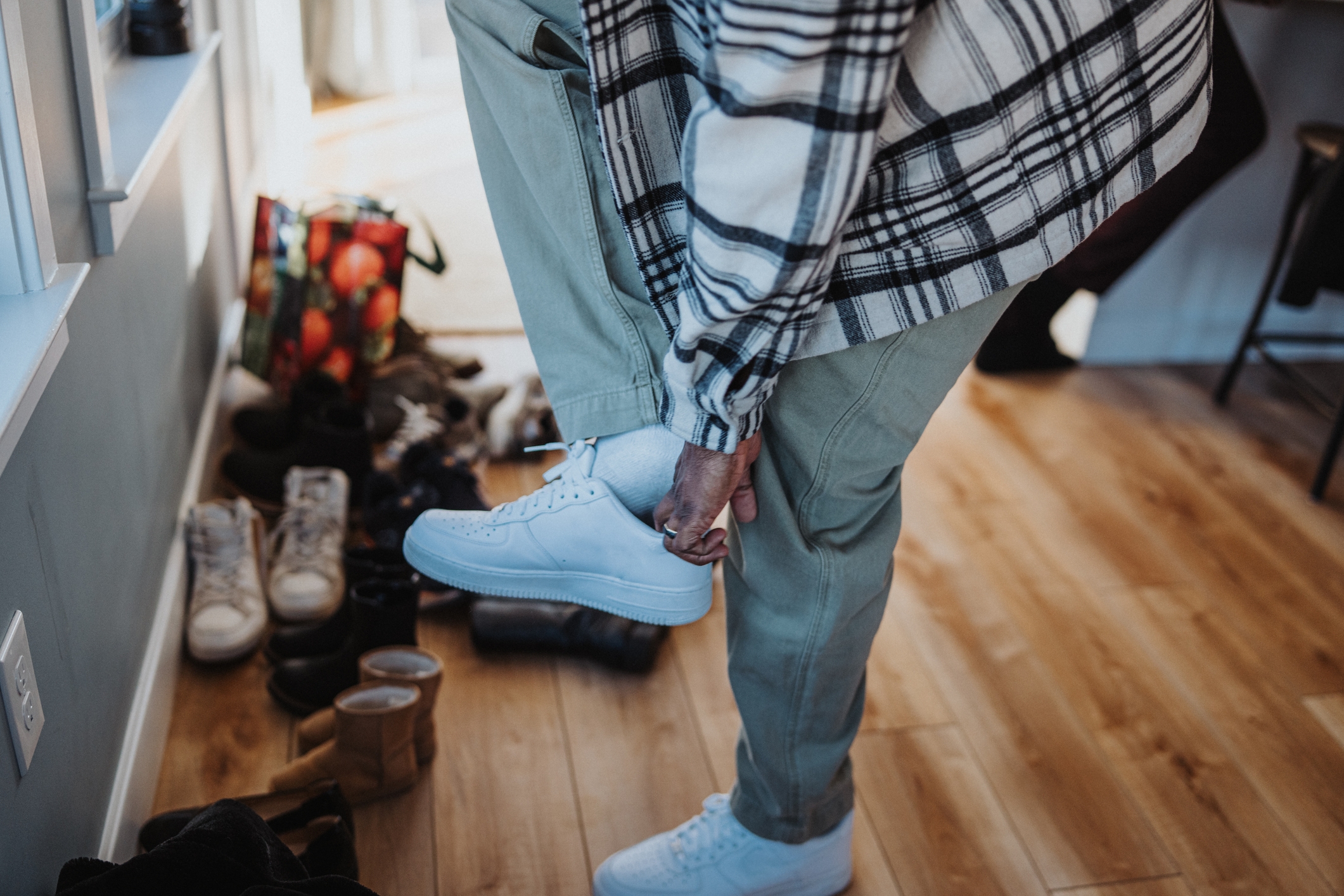 Person wearing a plaid shirt adjusts their sneaker in a room with scattered shoes