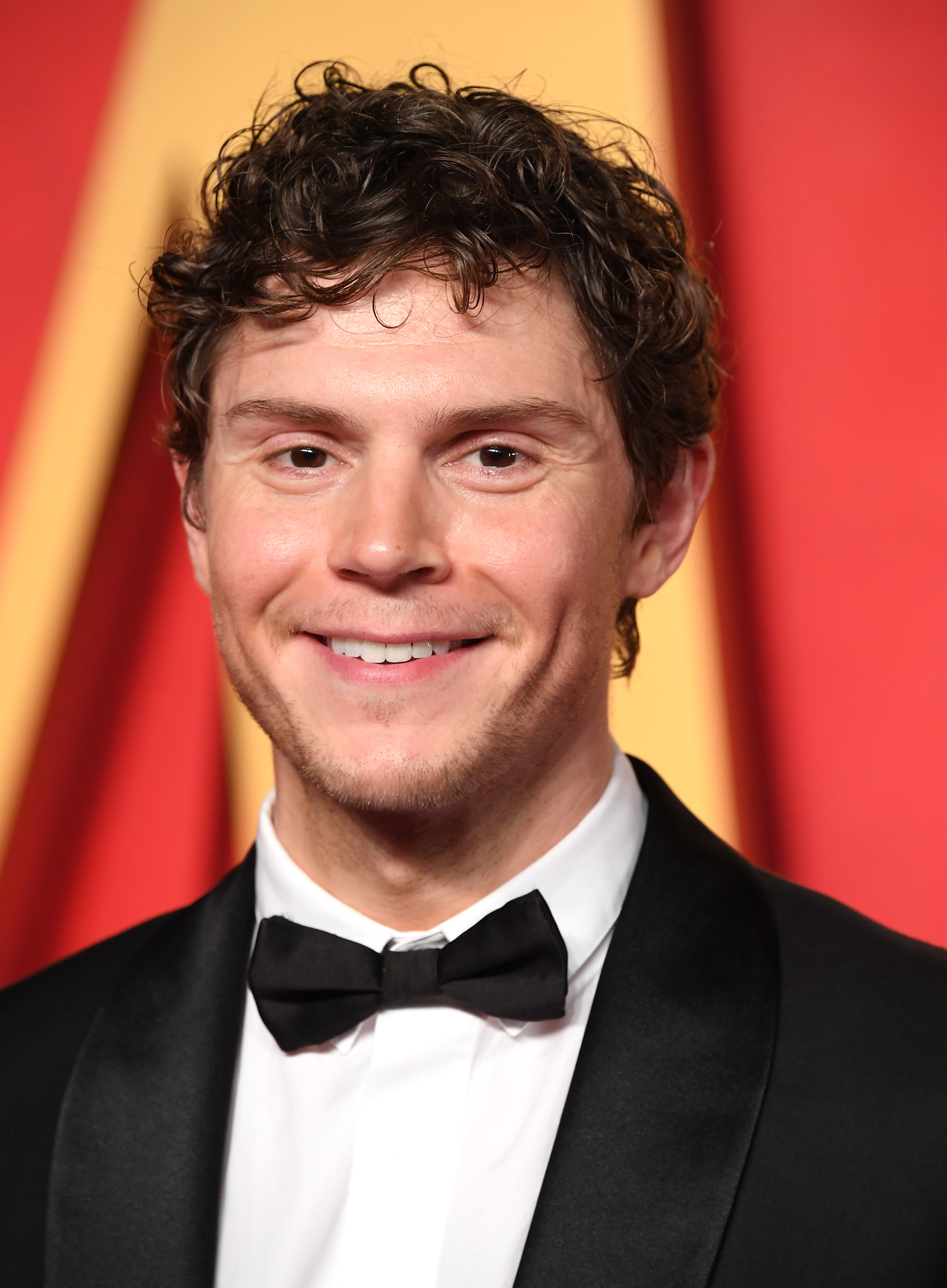 Person in formal attire with a black bow tie and suit, smiling on a red carpet event