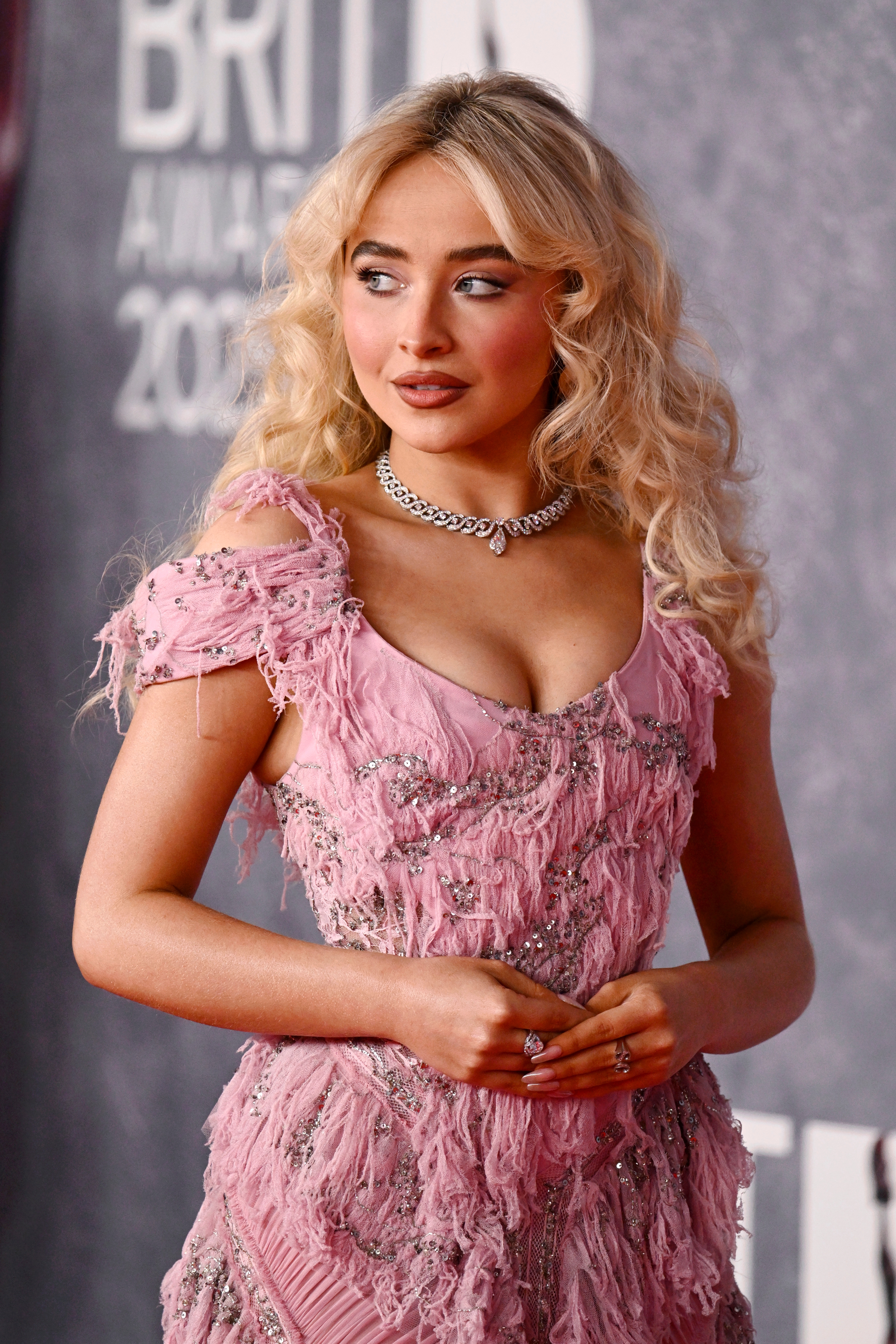 Person in an elegant, textured pink dress with off-the-shoulder sleeves and layered necklace, posing on a red carpet