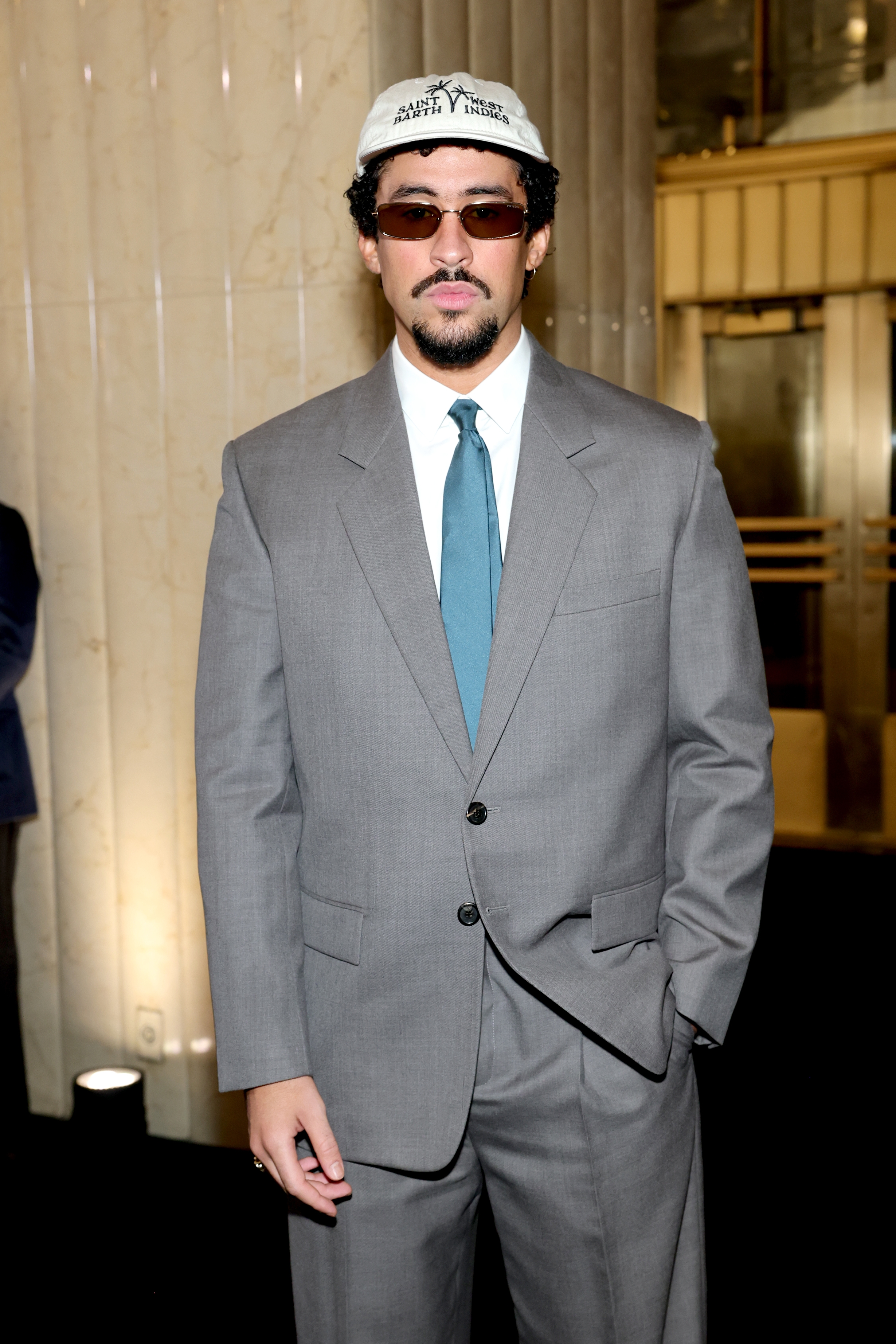 Person posing in a suit with sunglasses and a cap at an event, hands in pockets