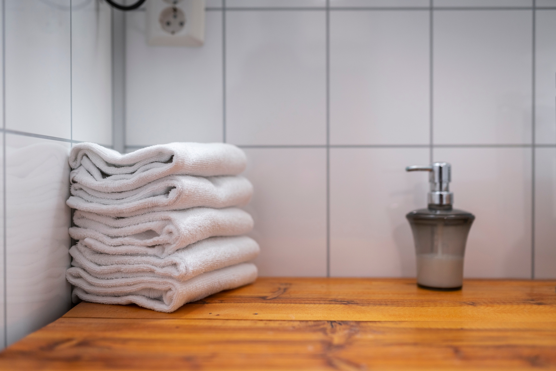 Folded towels stacked on a wooden counter next to a soap dispenser, set against a tiled wall background
