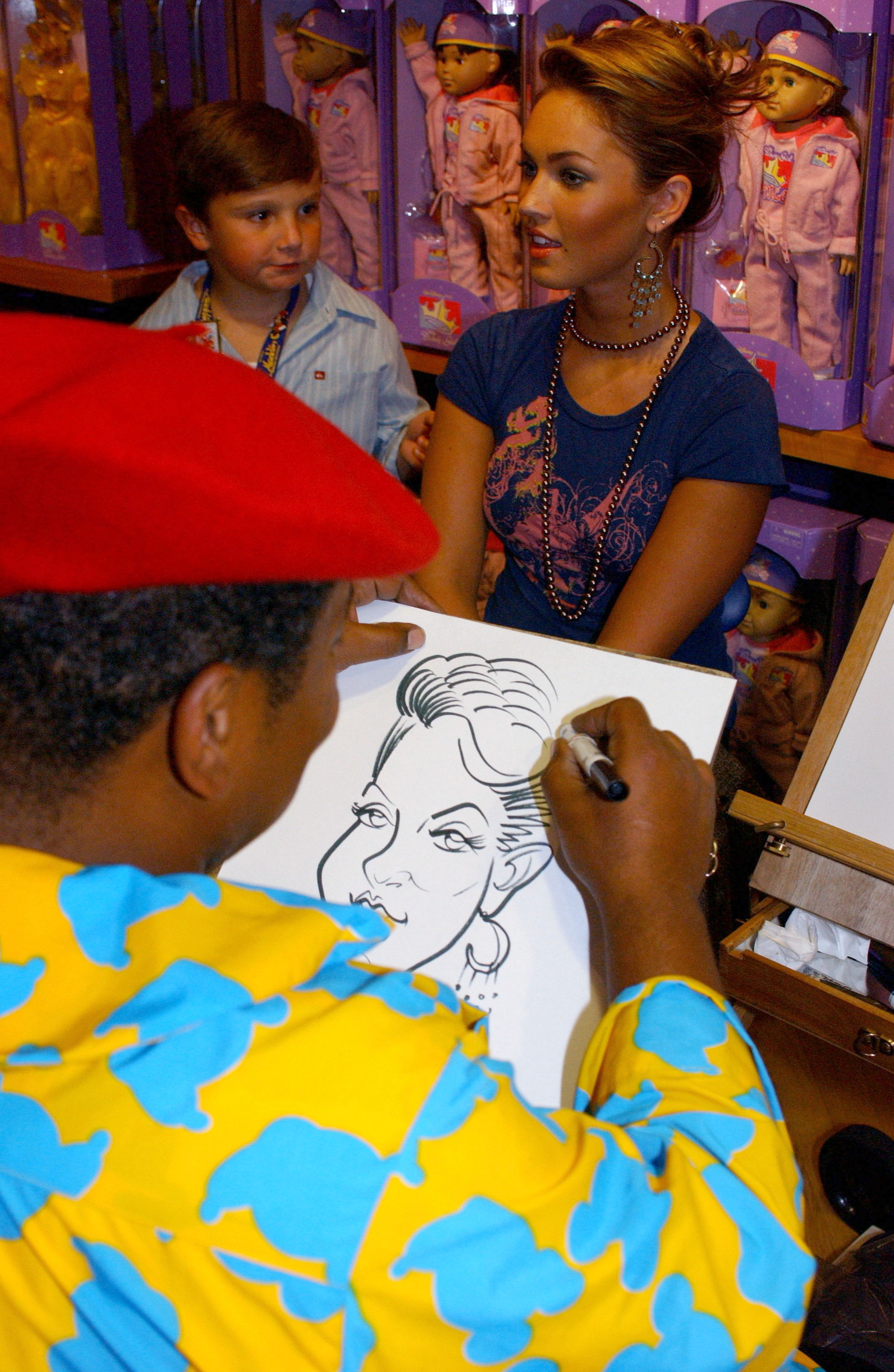 Artist sketches a caricature of a seated woman, while a boy watches nearby. Dolls displayed in background