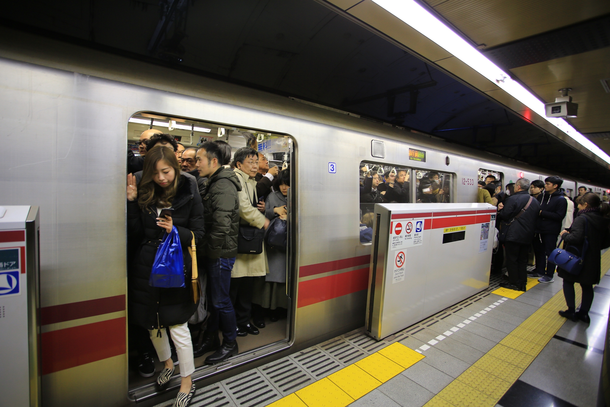 Crowded subway train with people tightly packed inside and on the platform, some using phones and others waiting for departure
