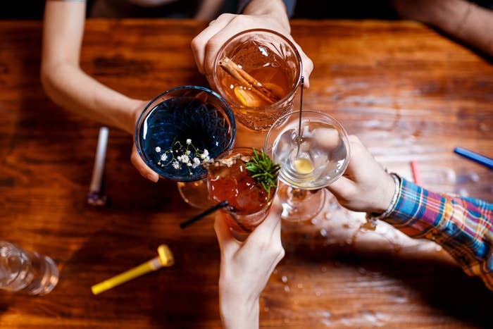 Four hands holding various cocktails in different glasses, toasting over a wooden table