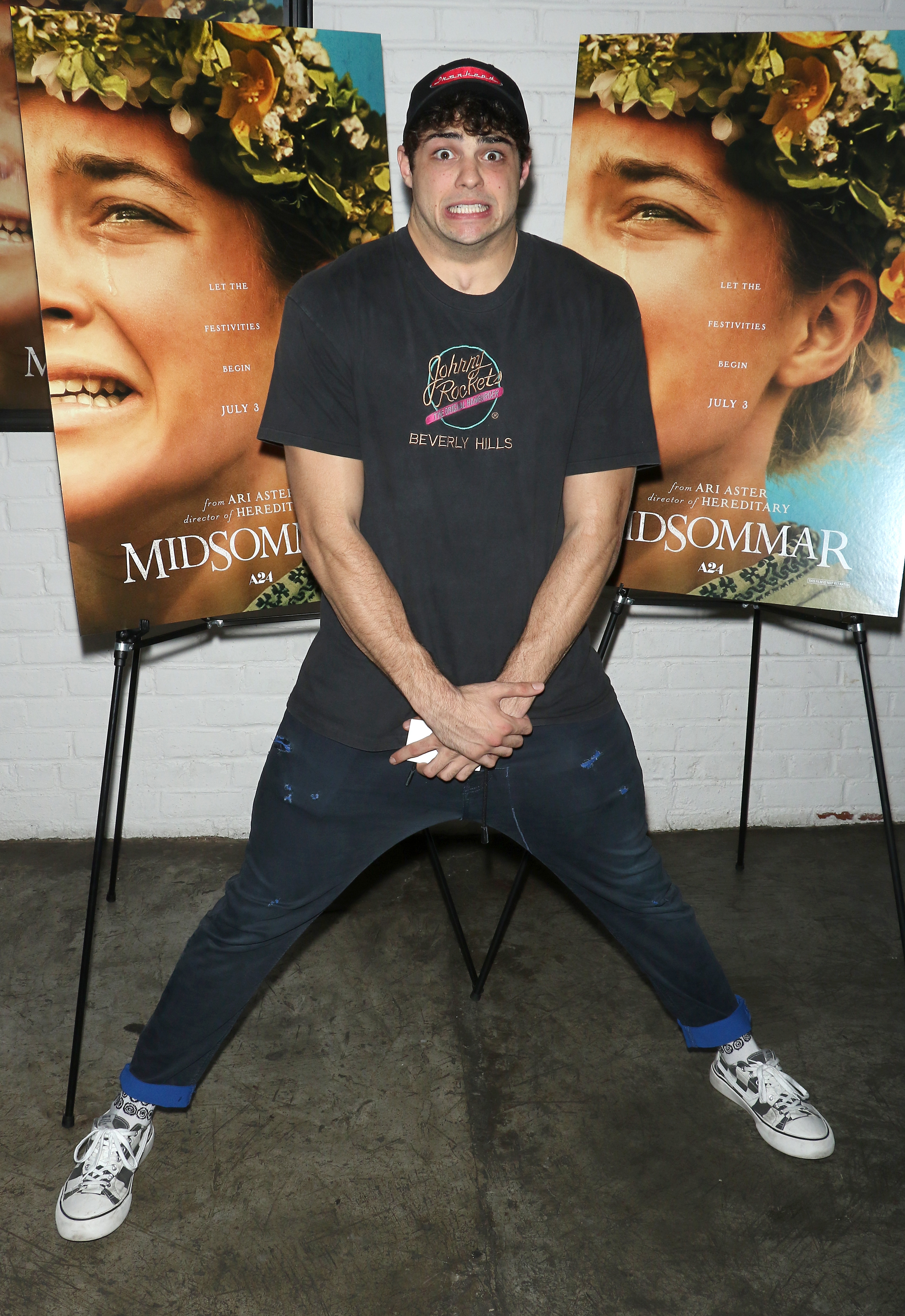 A person poses playfully with a nervous expression, standing in front of "Midsommar" movie posters, wearing casual streetwear and sneakers