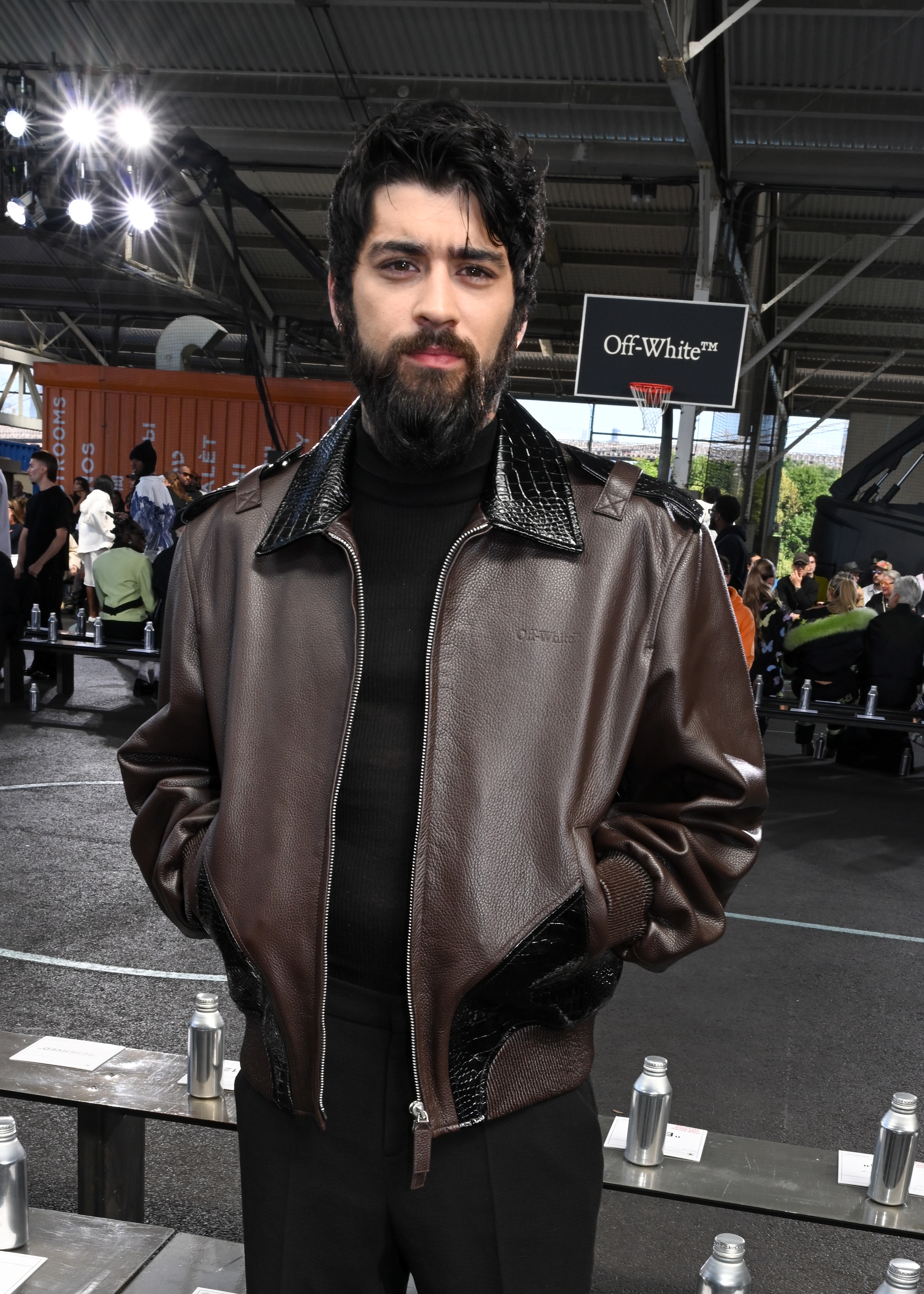 Person in a leather jacket and turtleneck at a fashion event, standing in an industrial-style venue with people seated in the background