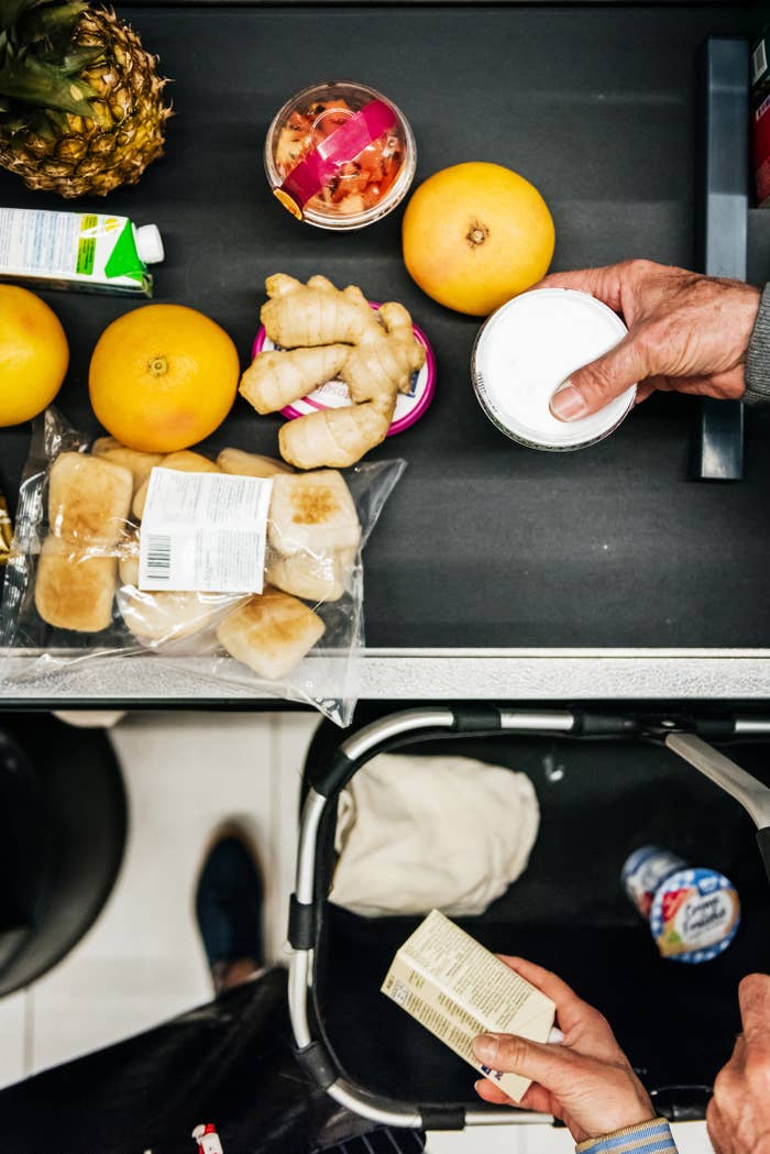 Grocery items, including grapefruits, ginger, and packaged goods, are placed on a checkout conveyor belt. Two hands hold items, preparing for purchase