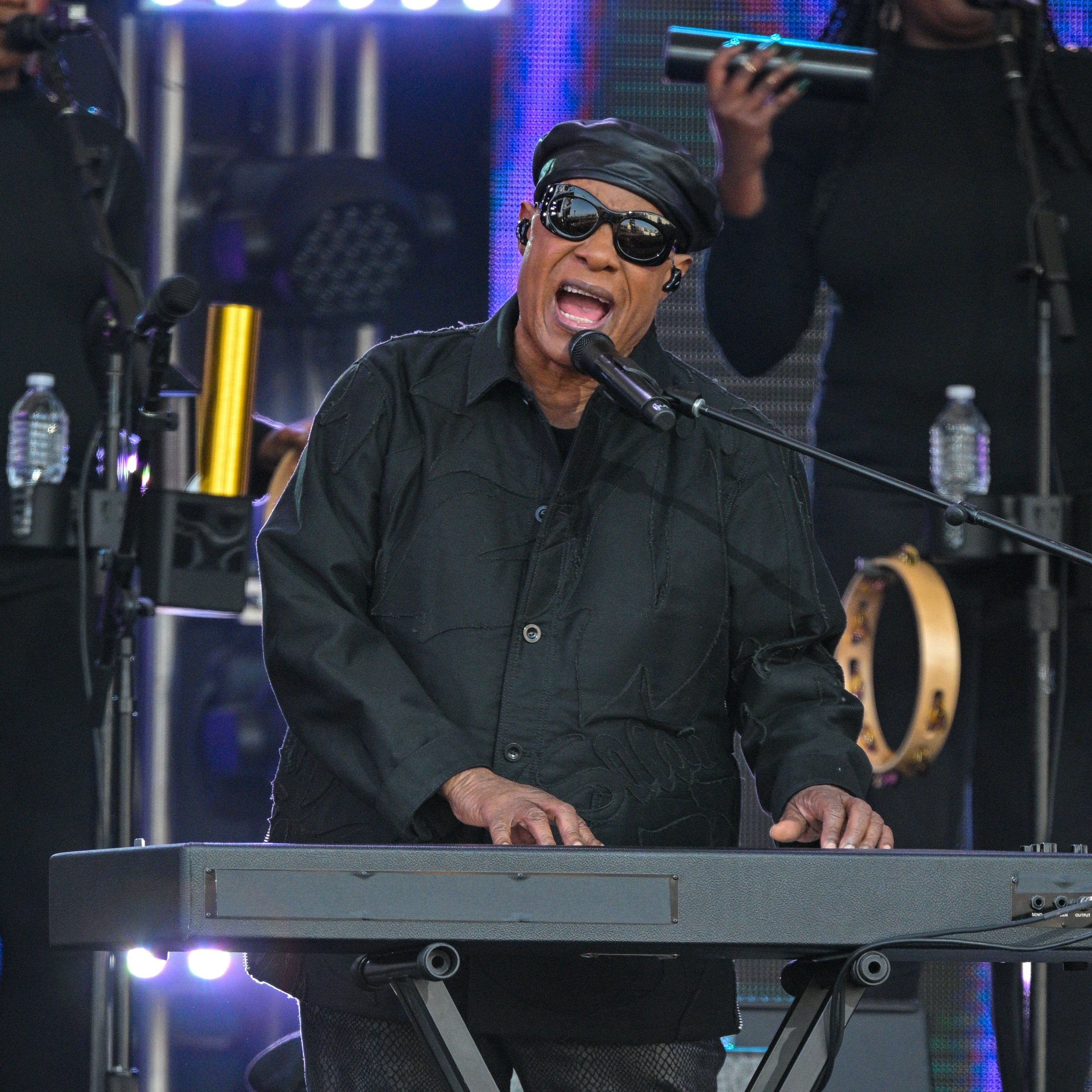 Musician performing energetically on stage, wearing dark sunglasses and a stylish hat, playing a keyboard. Background singers are visible