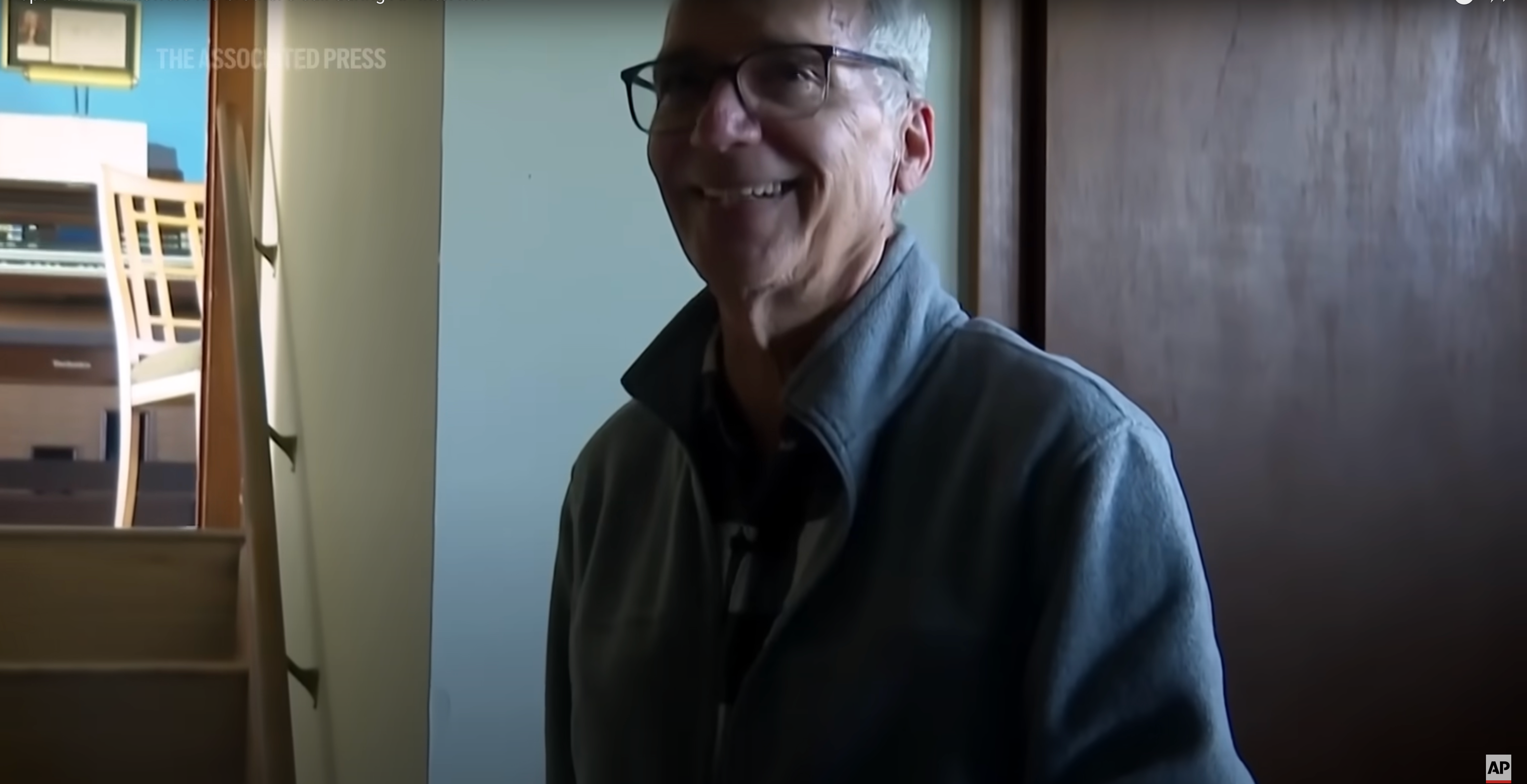 Person wearing glasses and a jacket, smiling indoors near a partially open door