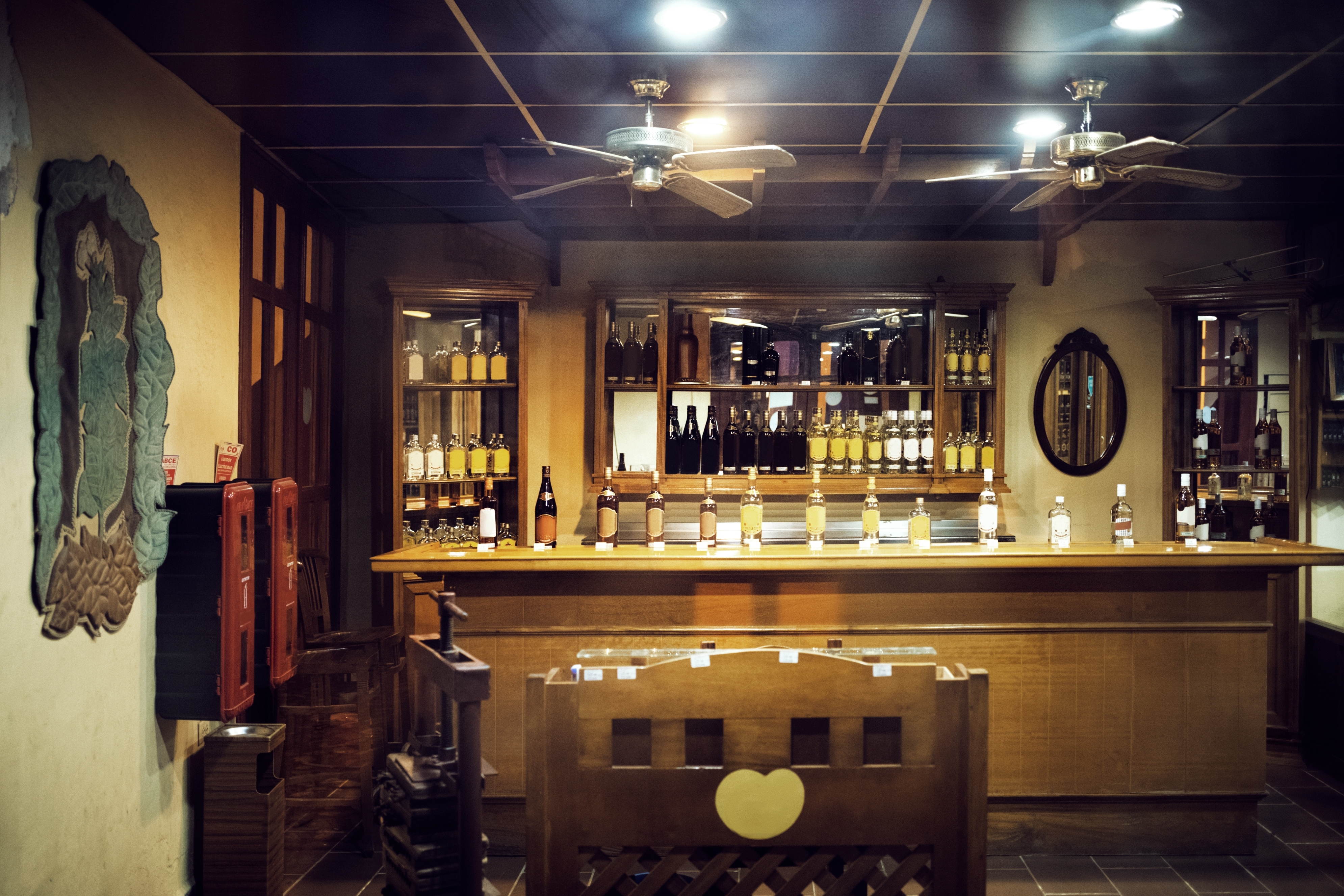 A cozy bar features a wooden counter lined with various bottles, shelves, and warm lighting, creating an inviting atmosphere for drinks