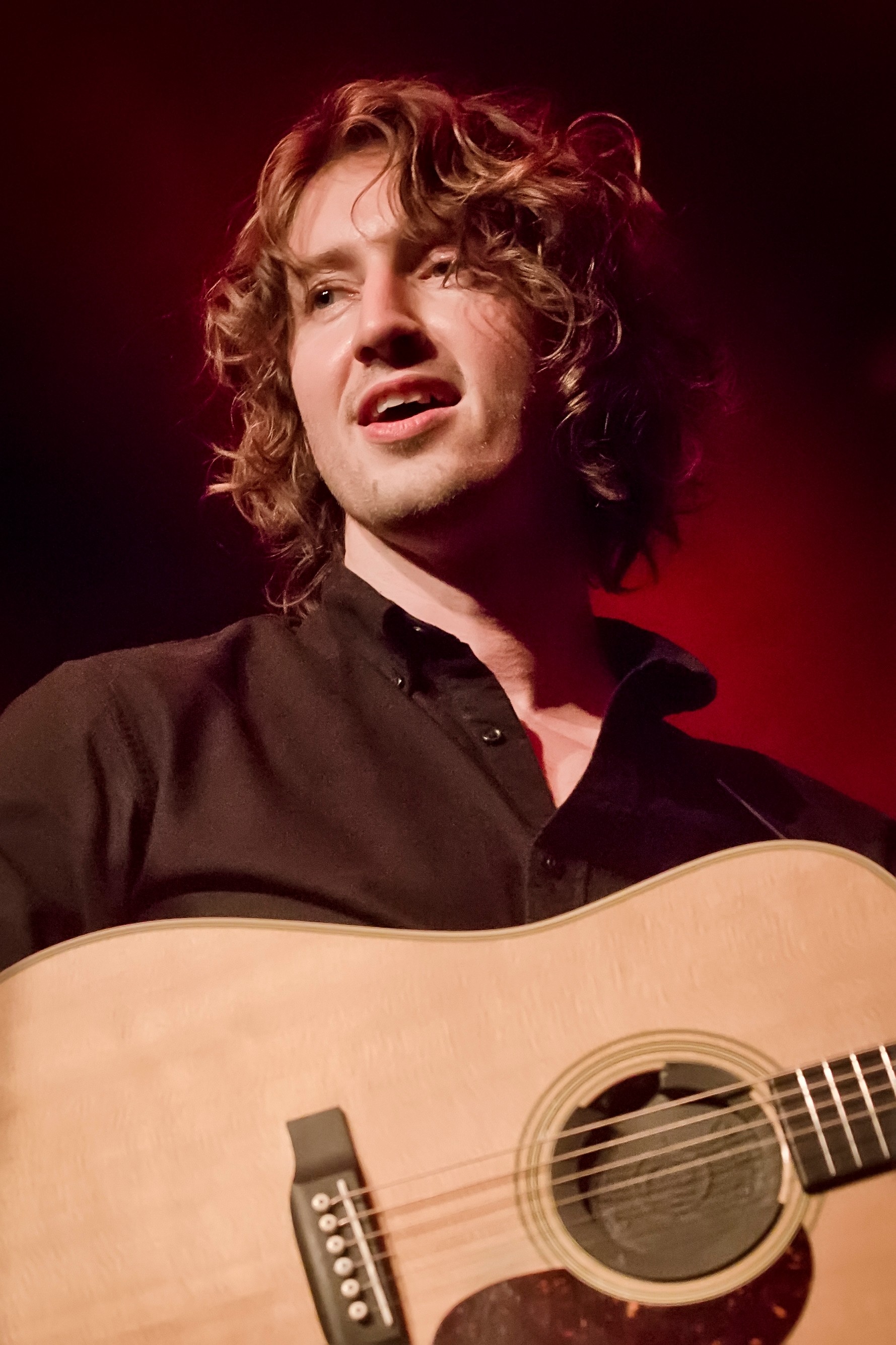Man performing on stage with acoustic guitar, wearing a dark shirt, against a blurred background
