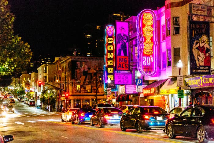 Street with neon signs for various clubs and venues, cars lined up on the side. Urban night scene with vibrant nightlife atmosphere