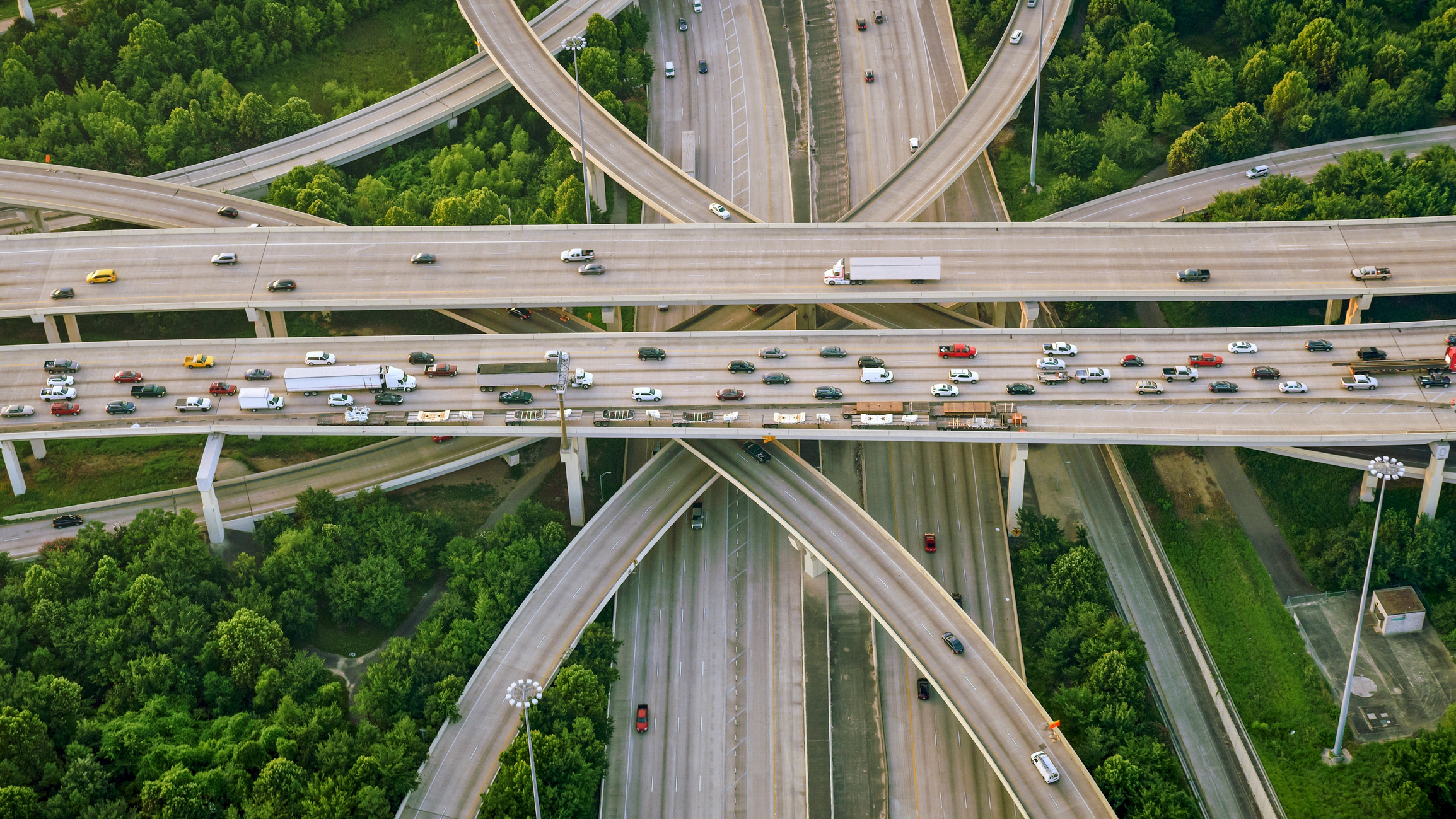 Aerial view of a complex highway interchange surrounded by green trees, with several vehicles traveling on the roads