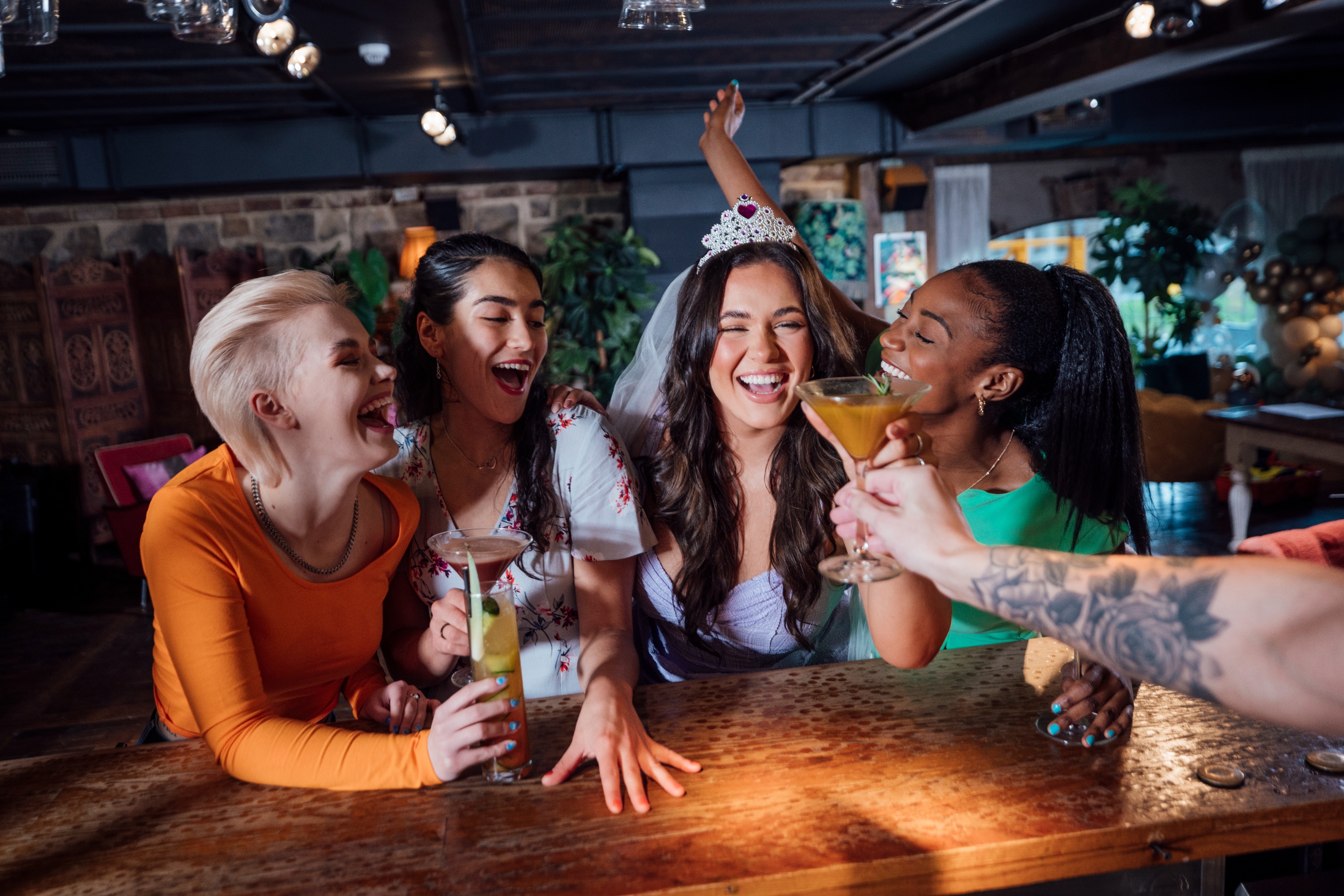 Group of friends at a bar, celebrating with drinks; one wears a tiara and veil, suggesting a bachelorette party