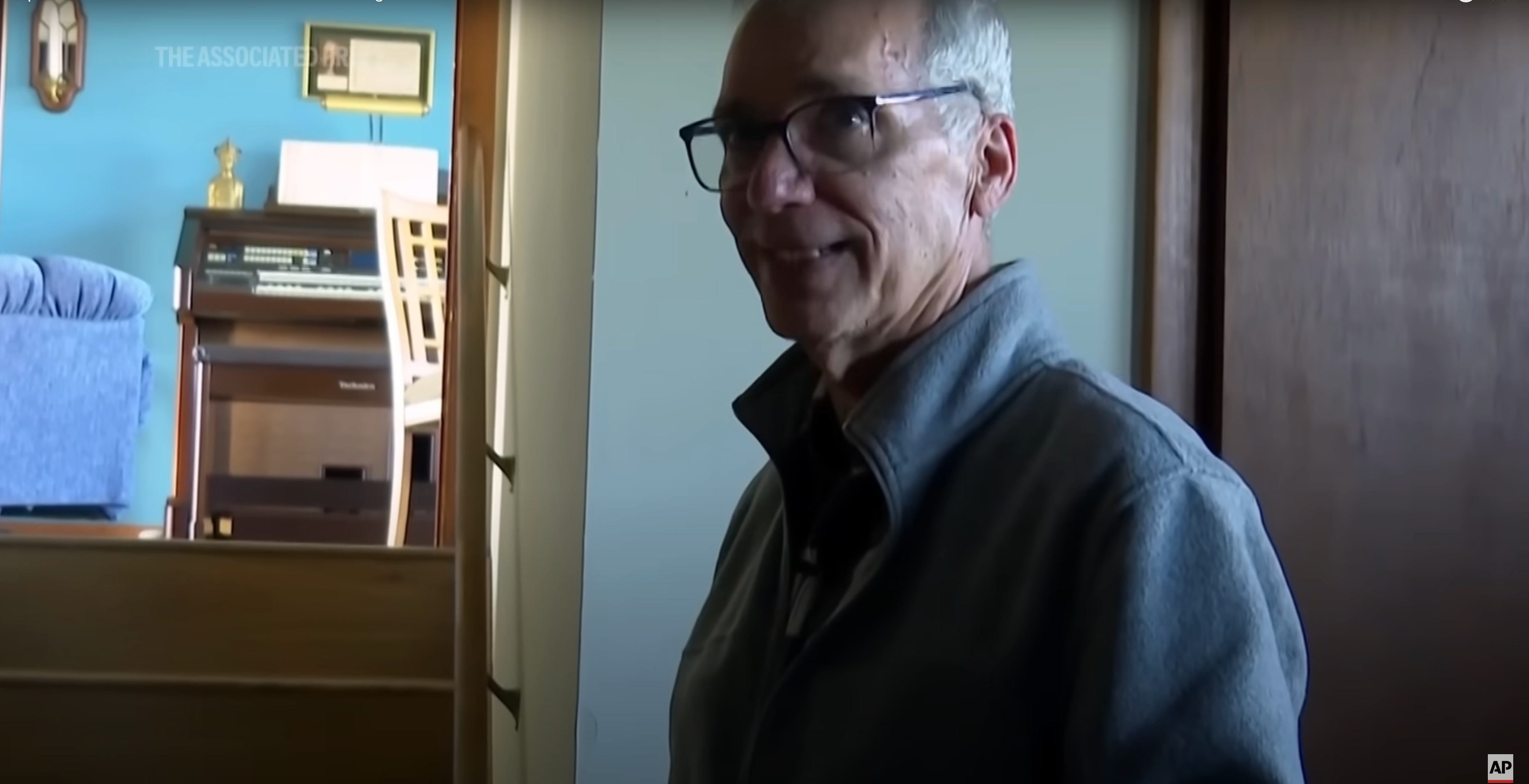 Man indoors near stairs, wearing glasses and a jacket, beside a room with a chair and an upright piano in the background
