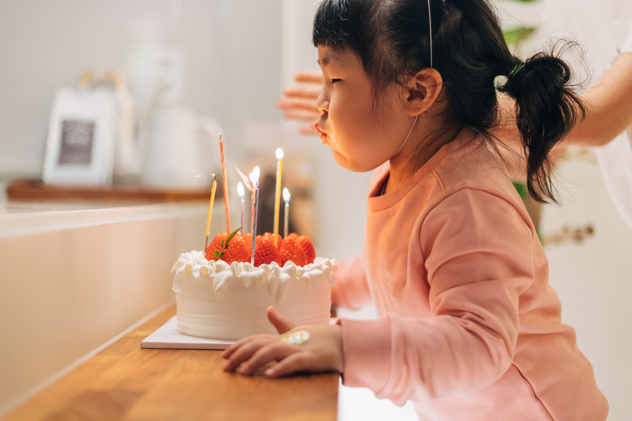 Child in a kitchen blowing out candles on a strawberry-topped cake, with an adult's hand supporting them gently