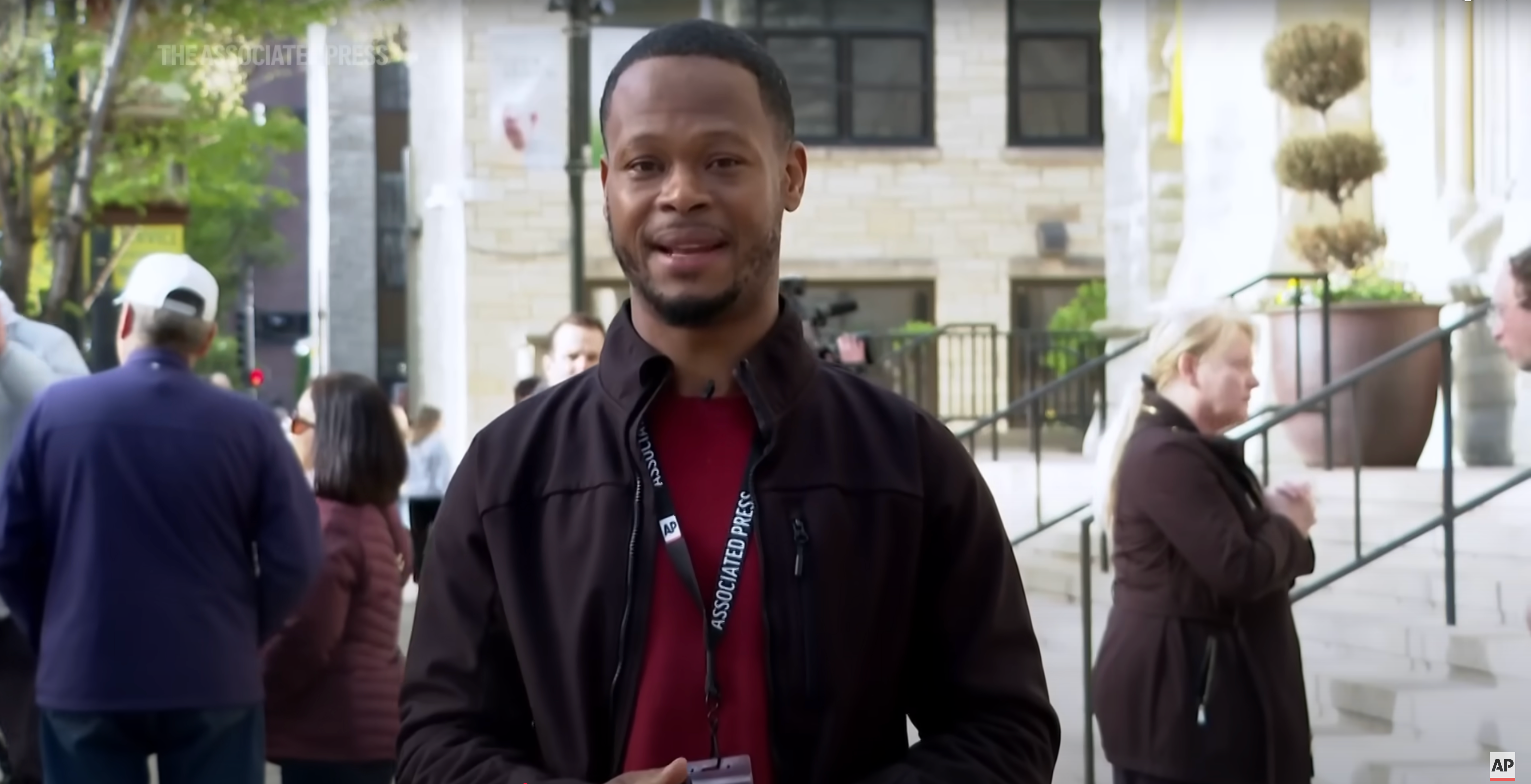 Man standing outdoors, wearing a black jacket and lanyard. People and buildings are in the background