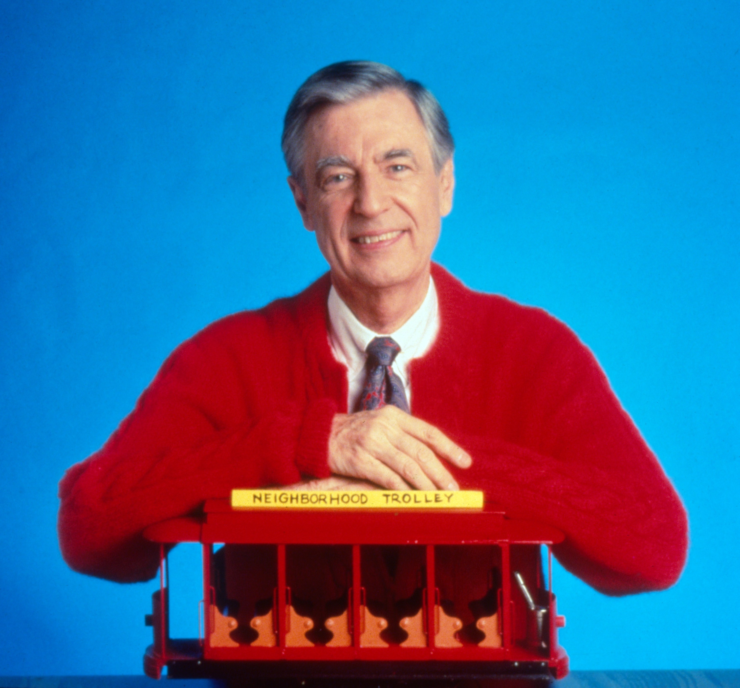 A person in a red cardigan smiles while holding a small model trolley labeled &quot;Neighborhood Trolley&quot; against a plain background