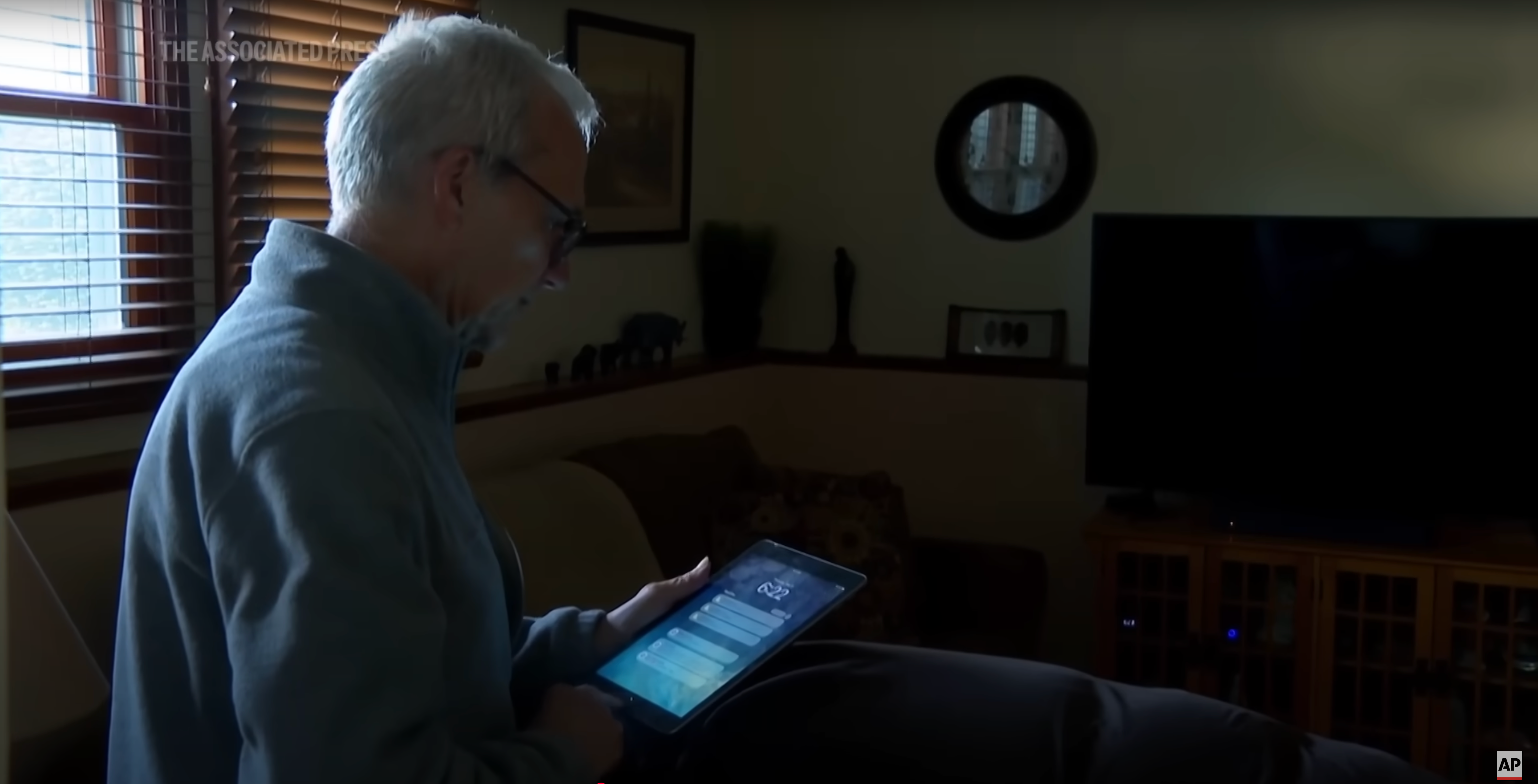An older adult using a tablet device in a dimly lit living room with a TV and wall art in the background