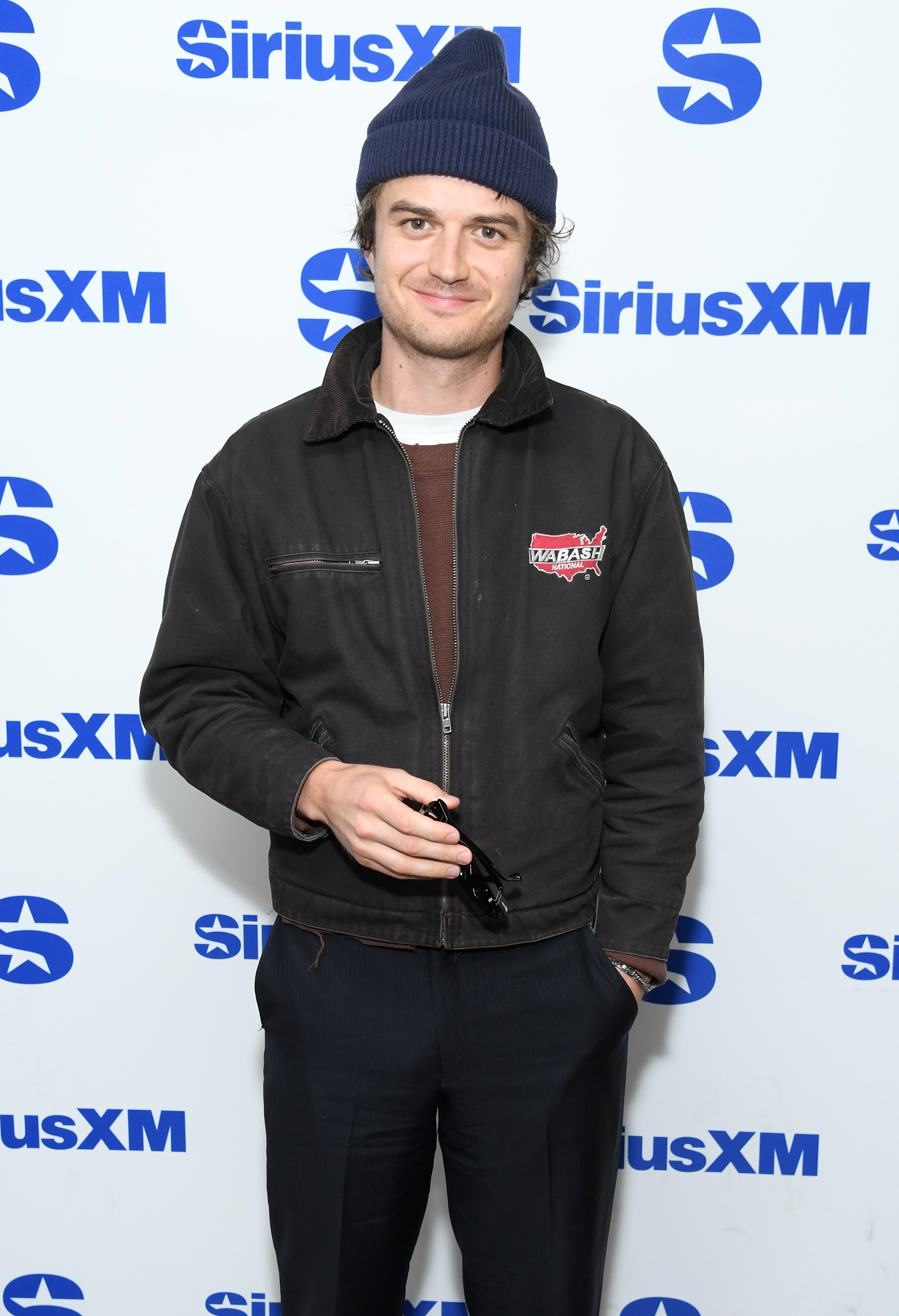 Person wearing a beanie and a casual jacket, holding sunglasses, poses in front of a SiriusXM backdrop at a media event
