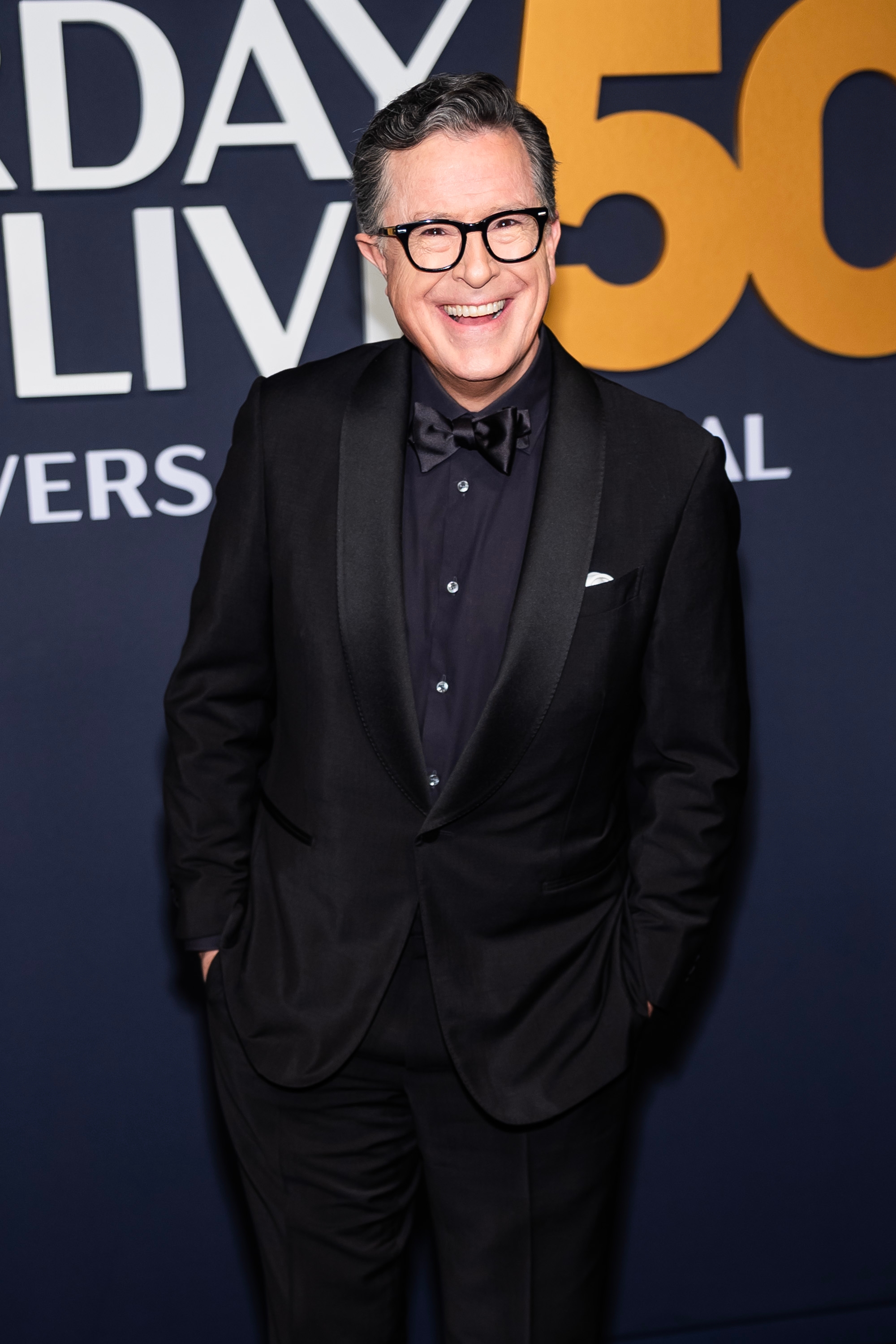 Person in a tuxedo smiles on a formal event red carpet with large text in the background