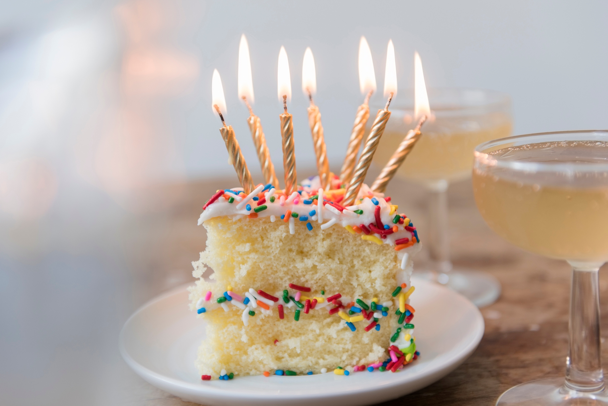 Slice of cake with lit candles and sprinkles, alongside two glasses of champagne