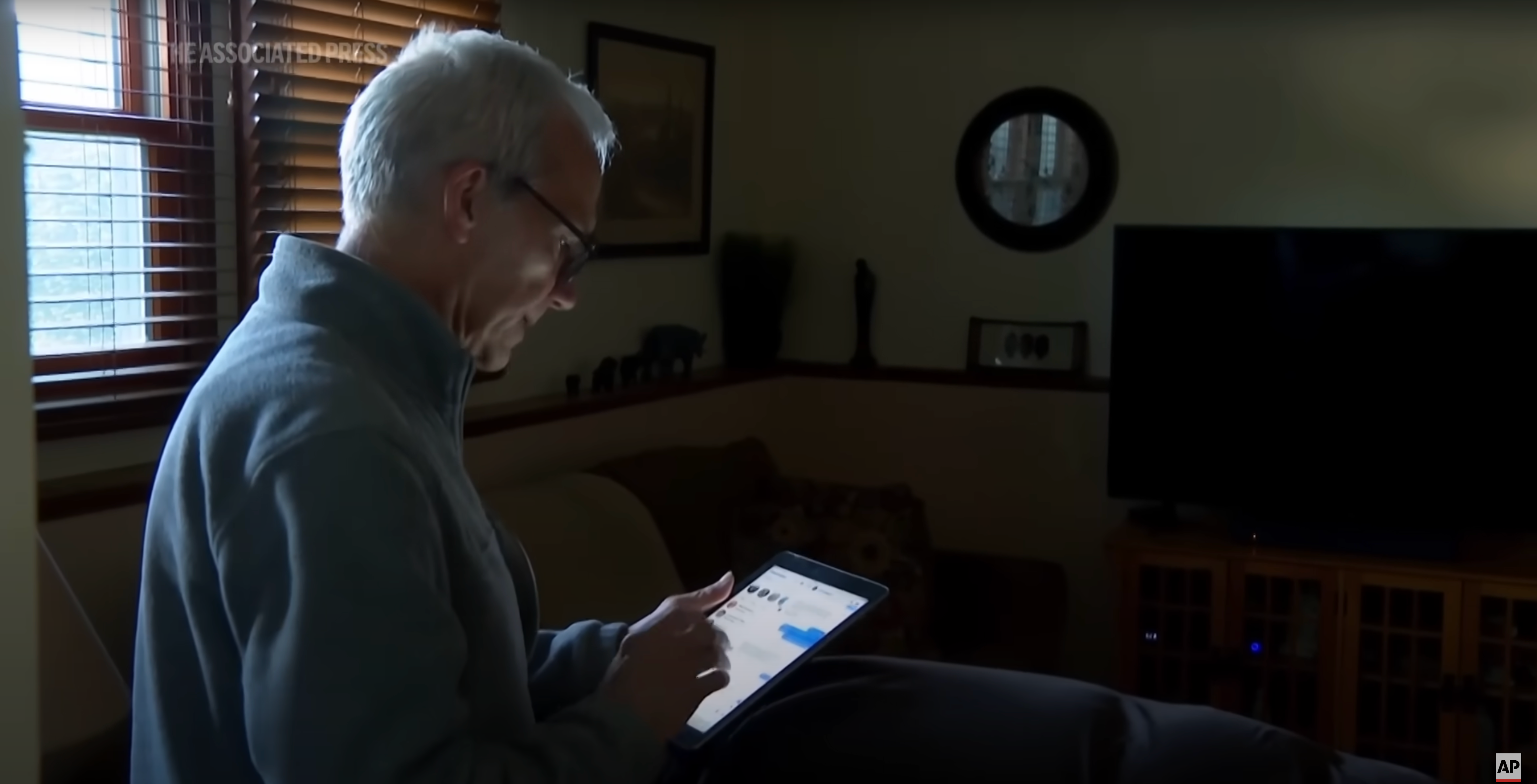 An older adult is using a tablet in a dimly lit living room with a window and a TV in the background